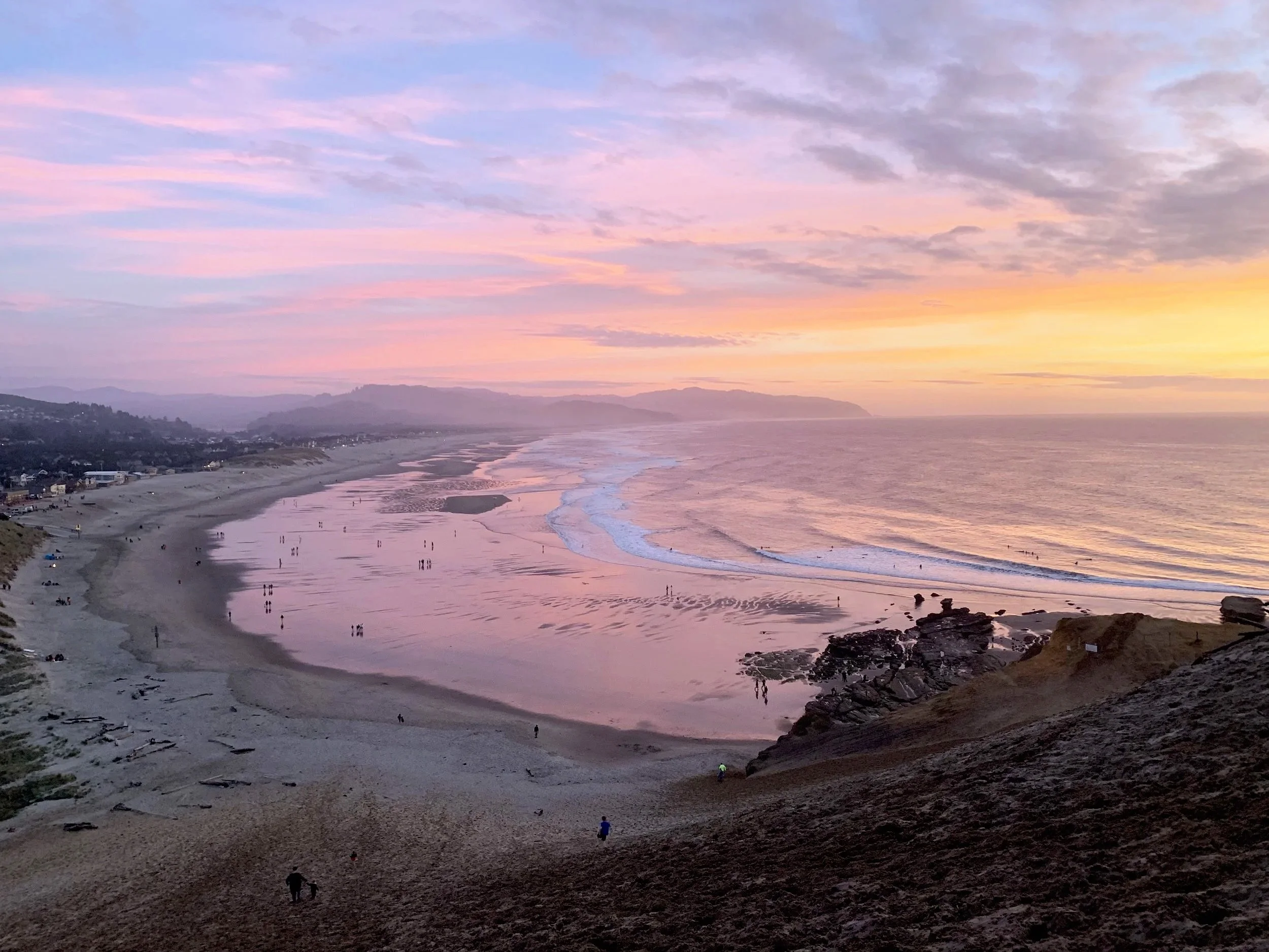 Sunset over a beach with pink and purple clouds, people walking along the shore, rocky outcroppings, and distant mountains.