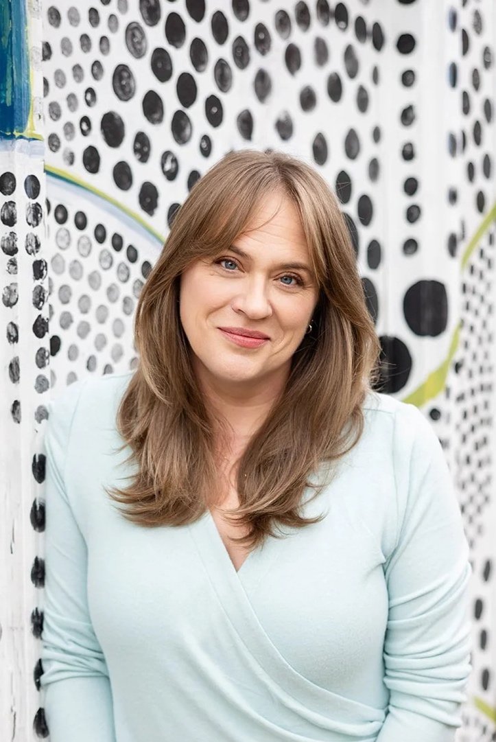 A woman with light brown hair and blue eyes wearing a light mint green top, standing in front of a black and white dotted wall.