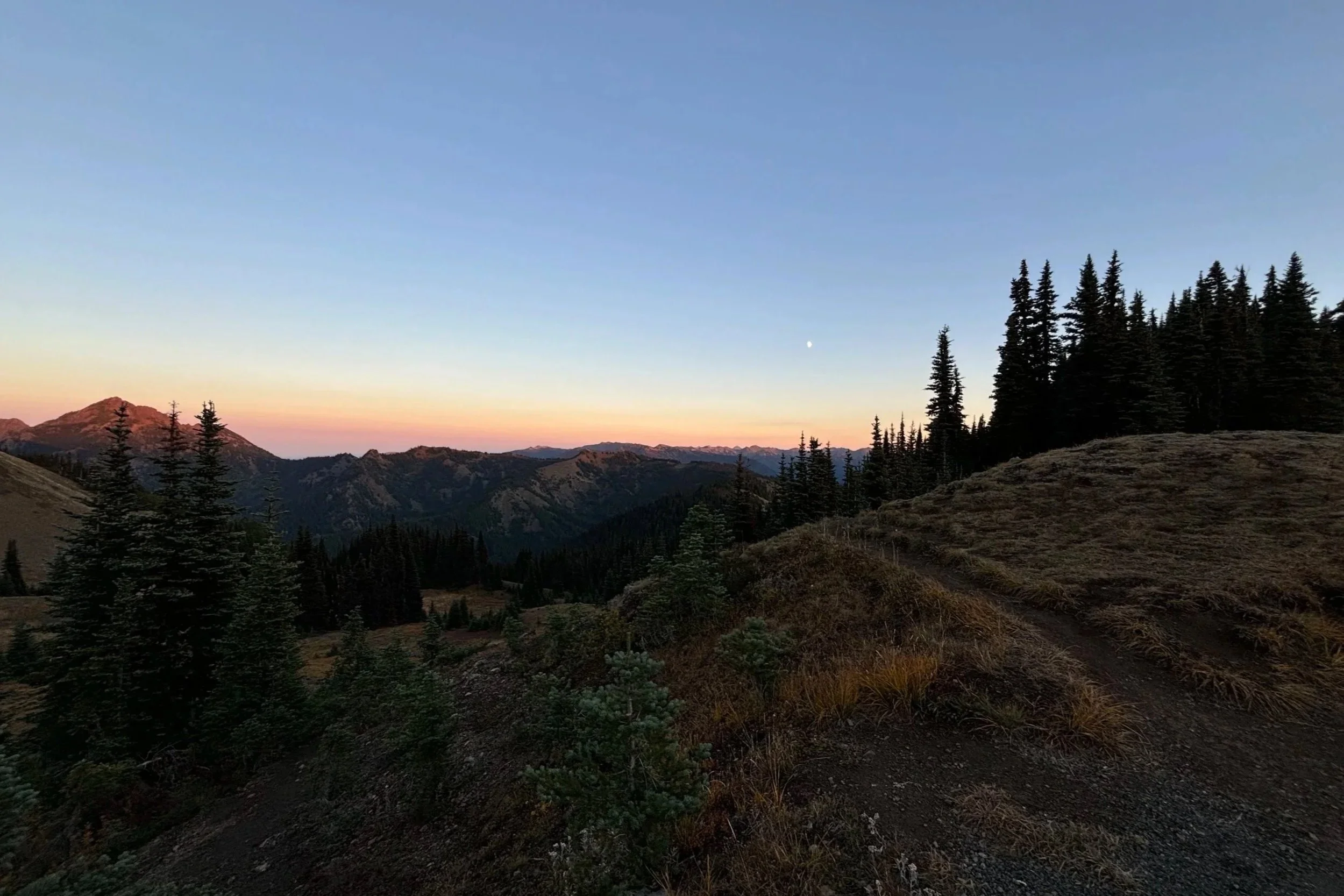 Scenic view of a mountain range during sunset with a clear sky, some trees, and a small moon visible in the distance.