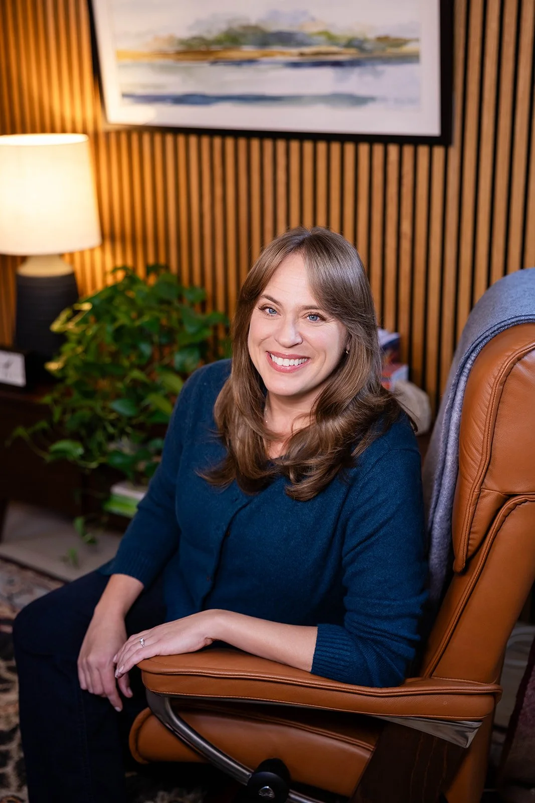 A woman with long brown hair, wearing a blue sweater, sitting on a brown leather chair in a warmly lit room with wooden panel walls, a lamp, a potted plant, and a painting in the background.