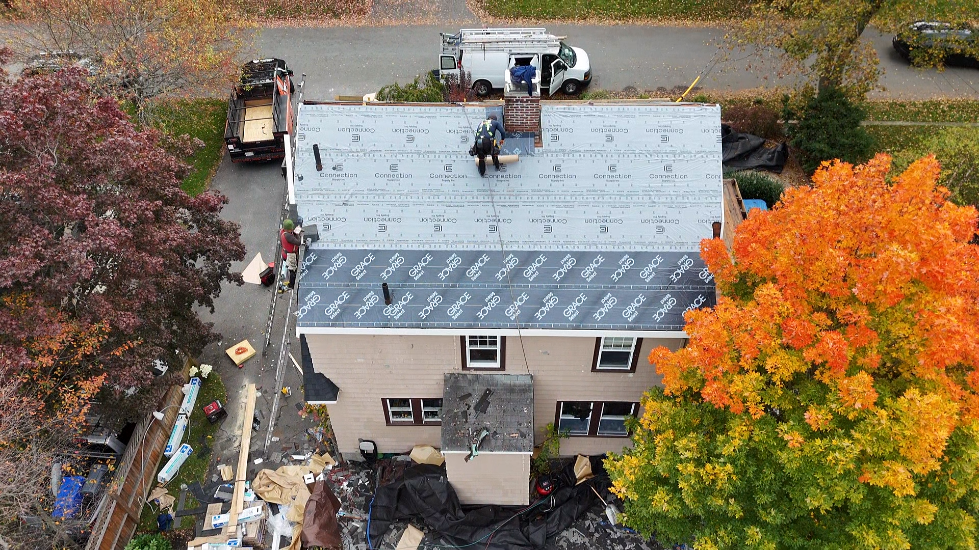 An aerial view of a two-story house under construction with workers installing roofing material. The house is surrounded by trees with autumn foliage.