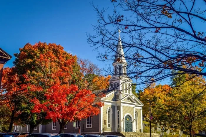 A white church with a tall steeple surrounded by colorful autumn trees with red, orange, and yellow leaves, under a clear blue sky.