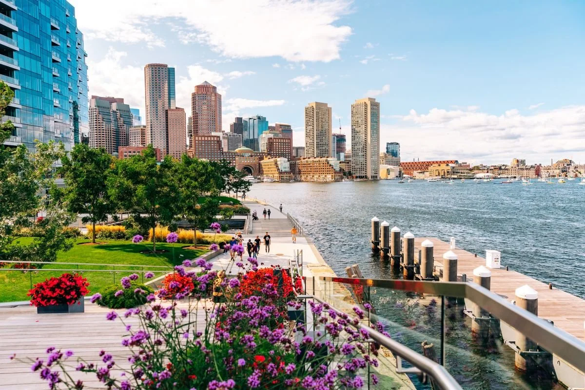 View of a city skyline across a body of water with a pier, trees, and colorful flowers in the foreground.