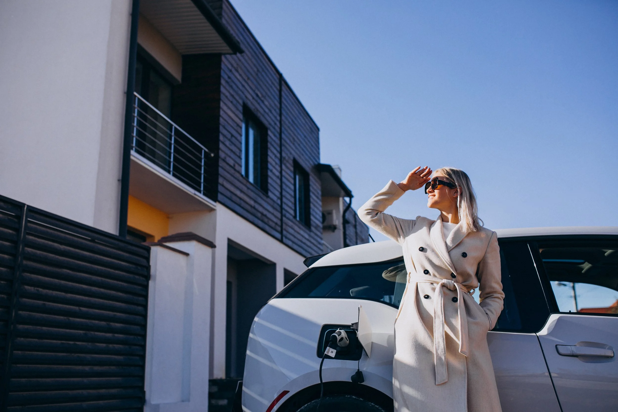 A woman in a beige trench coat and sunglasses standing beside a white electric car plugged into an charging station during daytime.