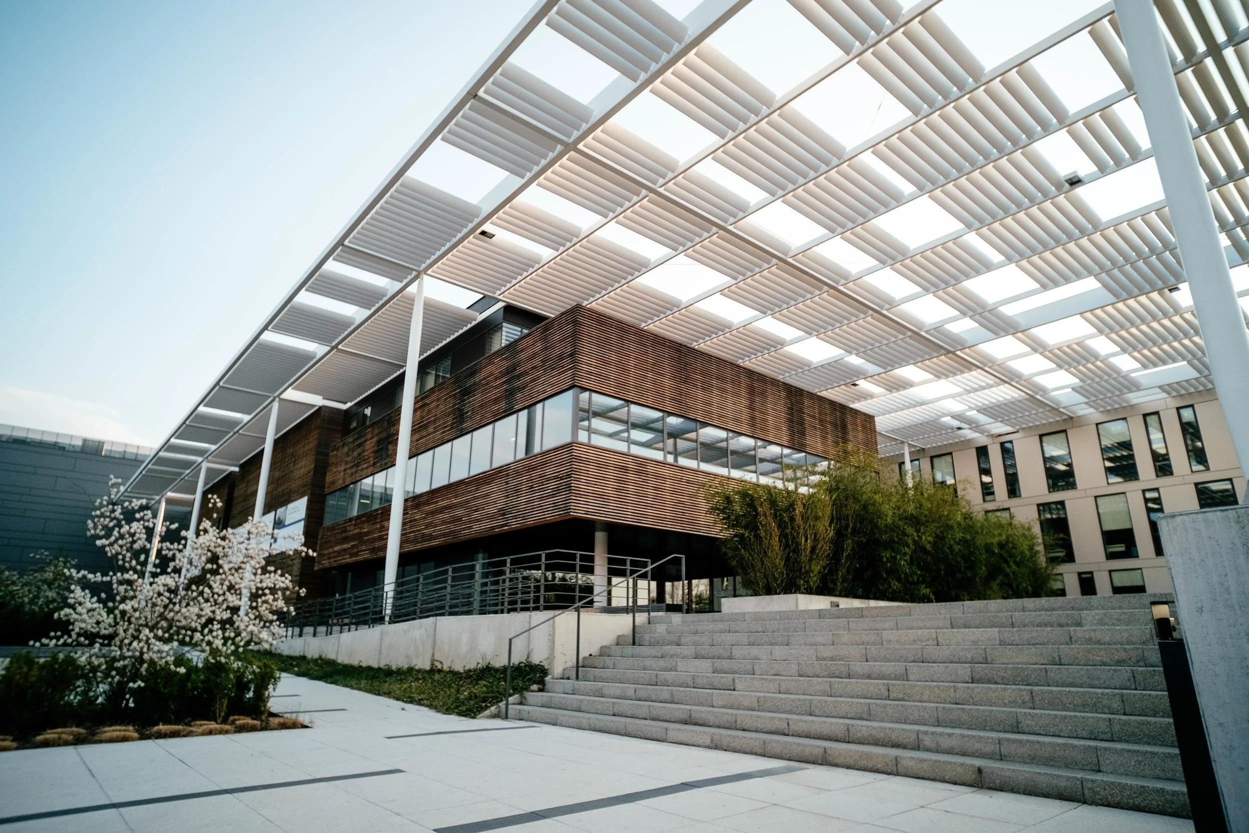 Modern building with wooden exterior and glass windows, surrounded by stairs, plants, and a unique white canopy structure overhead.