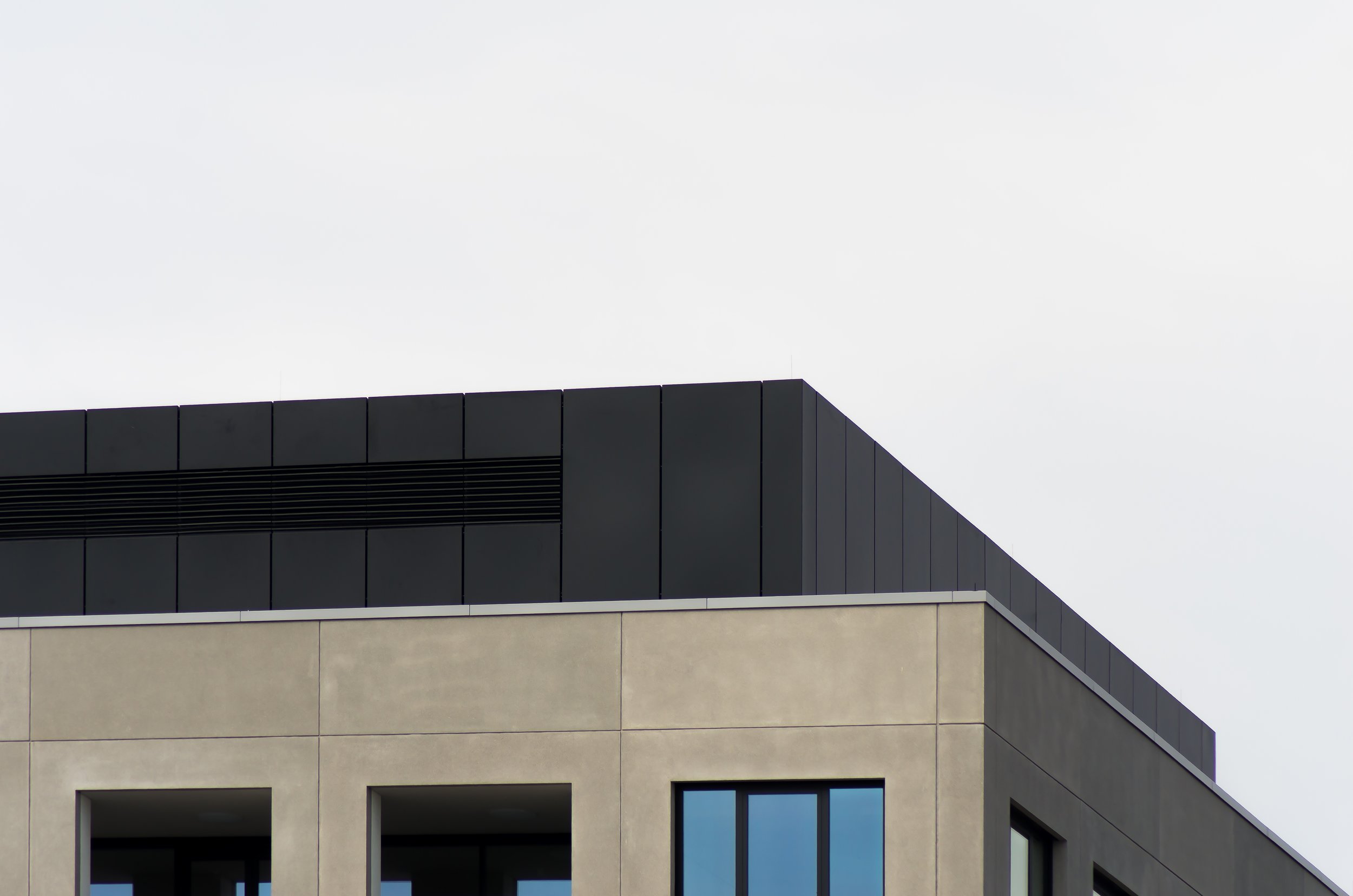 Close-up of the upper part of a modern building with beige and black exterior, featuring large windows and a grey overcast sky in the background.