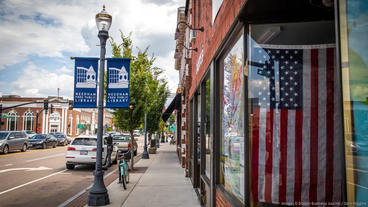 Street scene in Needham, Massachusetts with a row of brick buildings, parked cars, a lamppost with banners that read "Needham Free Public Library," and an American flag inside a store window.