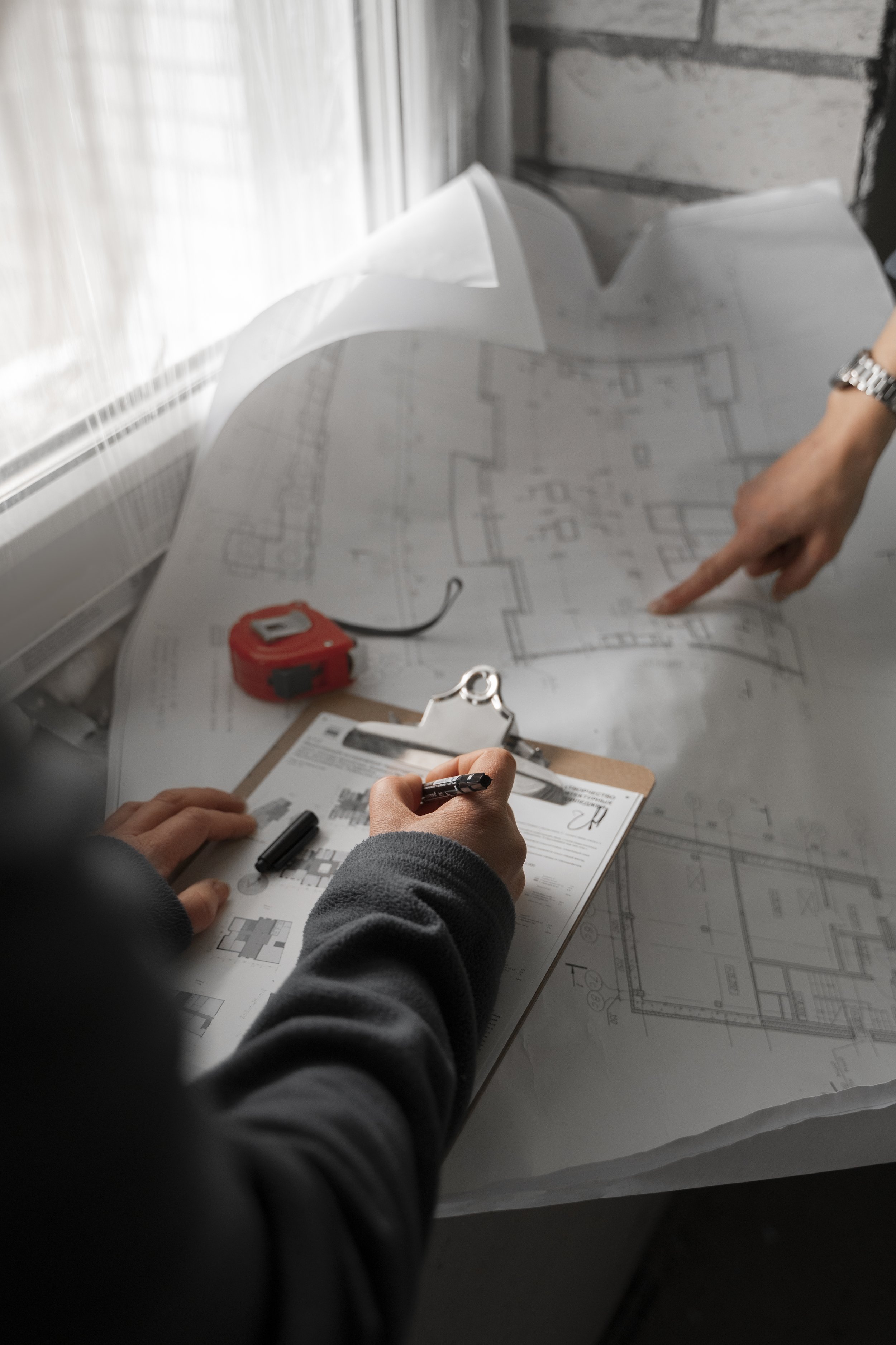 Two people reviewing and pointing at an architectural blueprint on a table, with a clipboard, pen, and measuring tape nearby, near a window with natural light.