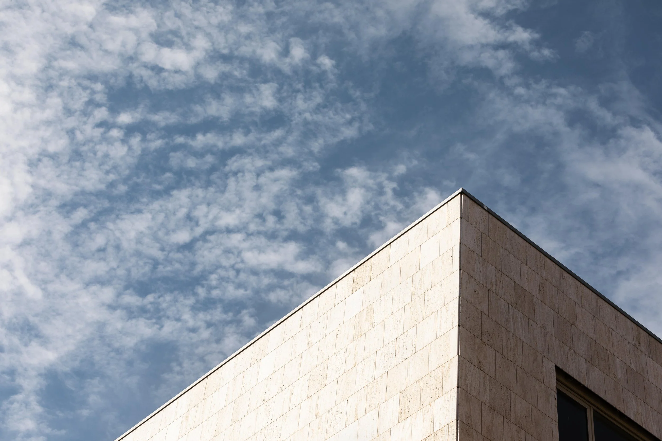 Corner of a modern building with beige stone facade against a blue sky with scattered clouds.