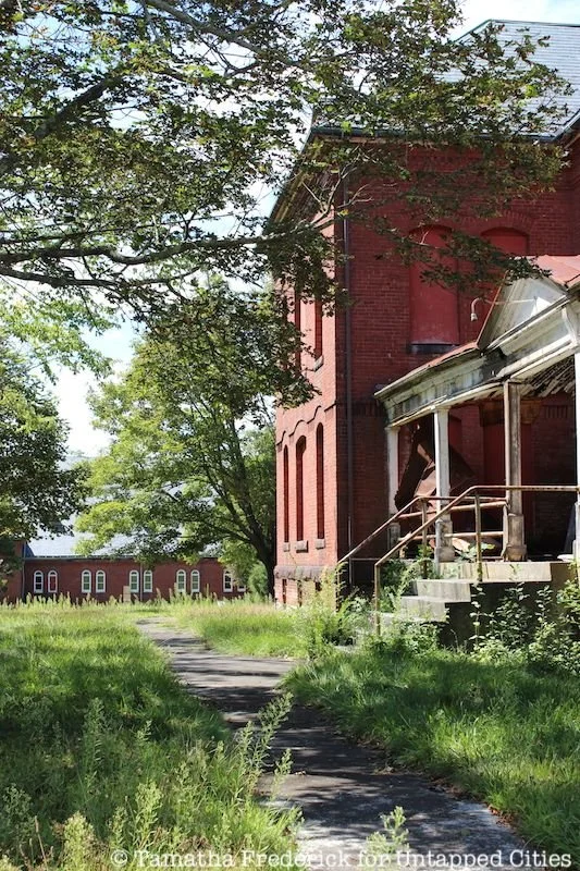 An old red brick building with a porch, stairs, and a metal railing, surrounded by overgrown grass and trees, on a sunny day.