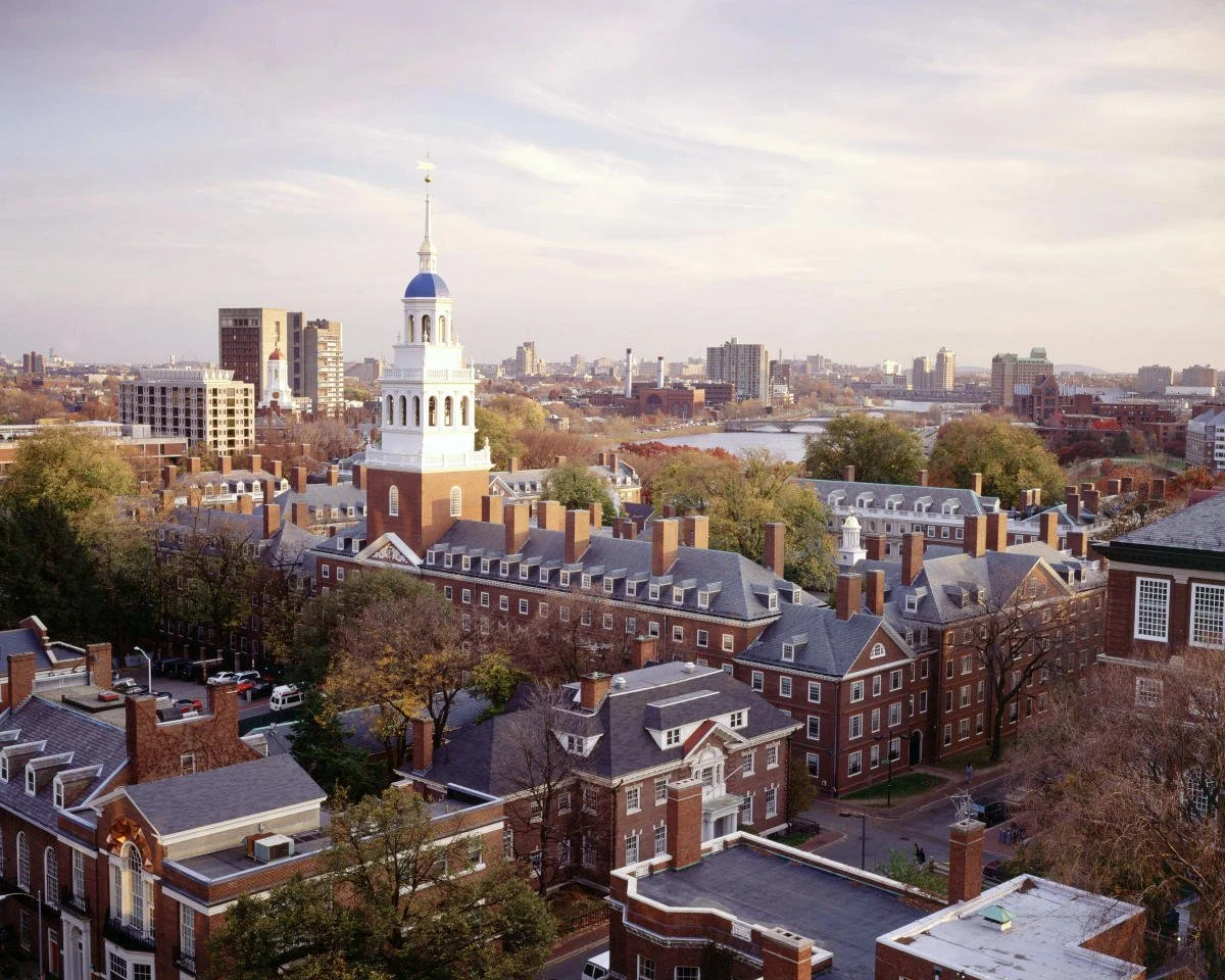 An aerial view of historic campus buildings with a city skyline and river in the background during daytime.