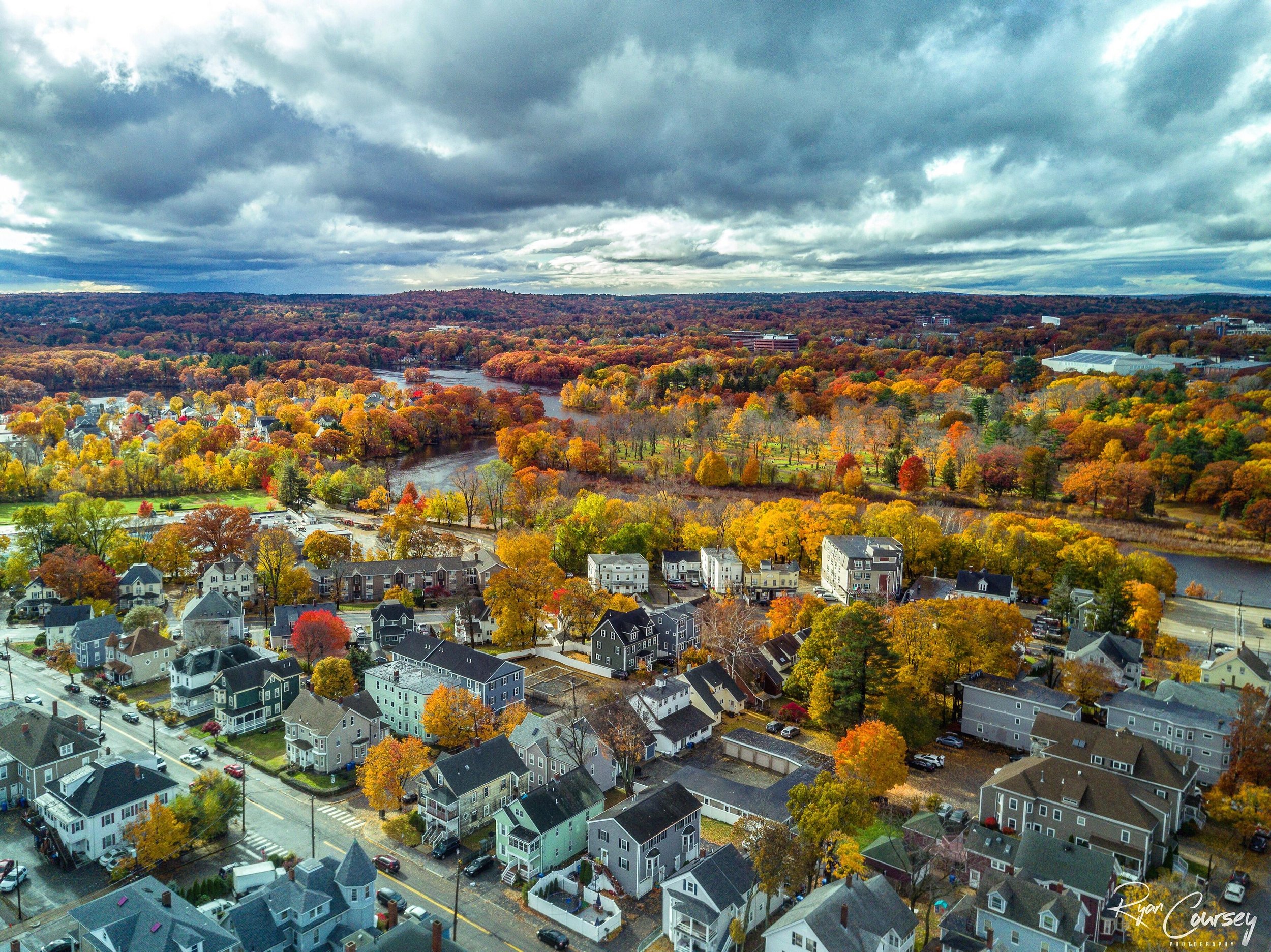 An aerial view of a neighborhood surrounded by a river and a forest with autumn foliage, under a cloudy sky.
