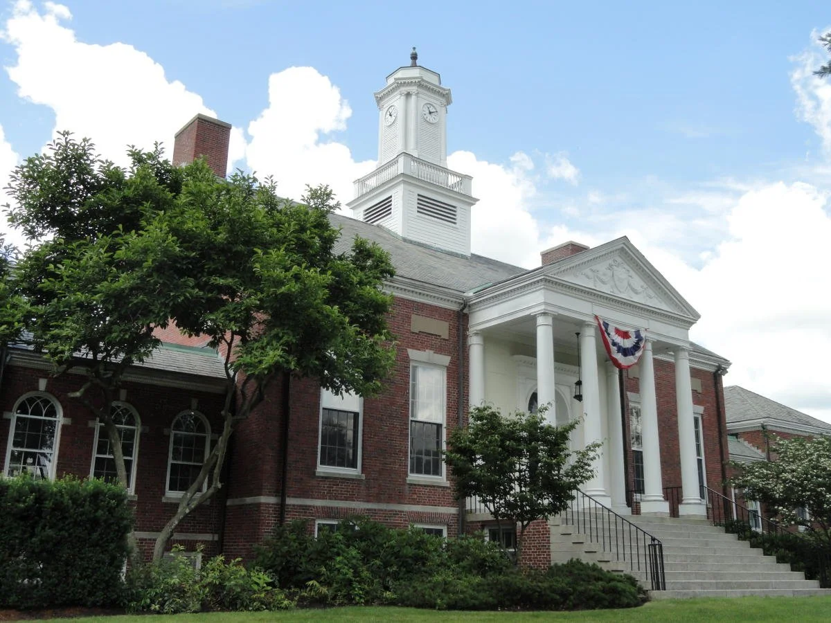 A historic brick building with white columns and a clock tower, decorated with American patriotic bunting, surrounded by trees and greenery under a partly cloudy sky.