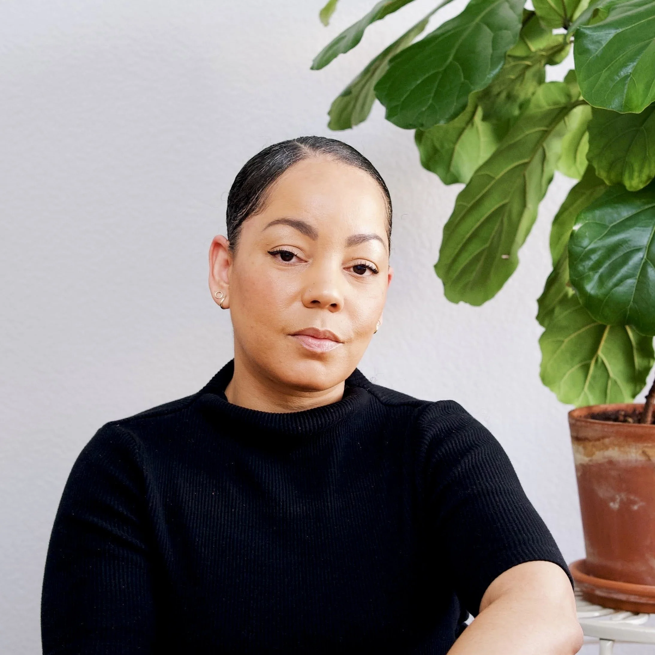A woman with dark hair pulled back, wearing a black top, sitting beside a large green plant in a terracotta pot, against a plain white wall.