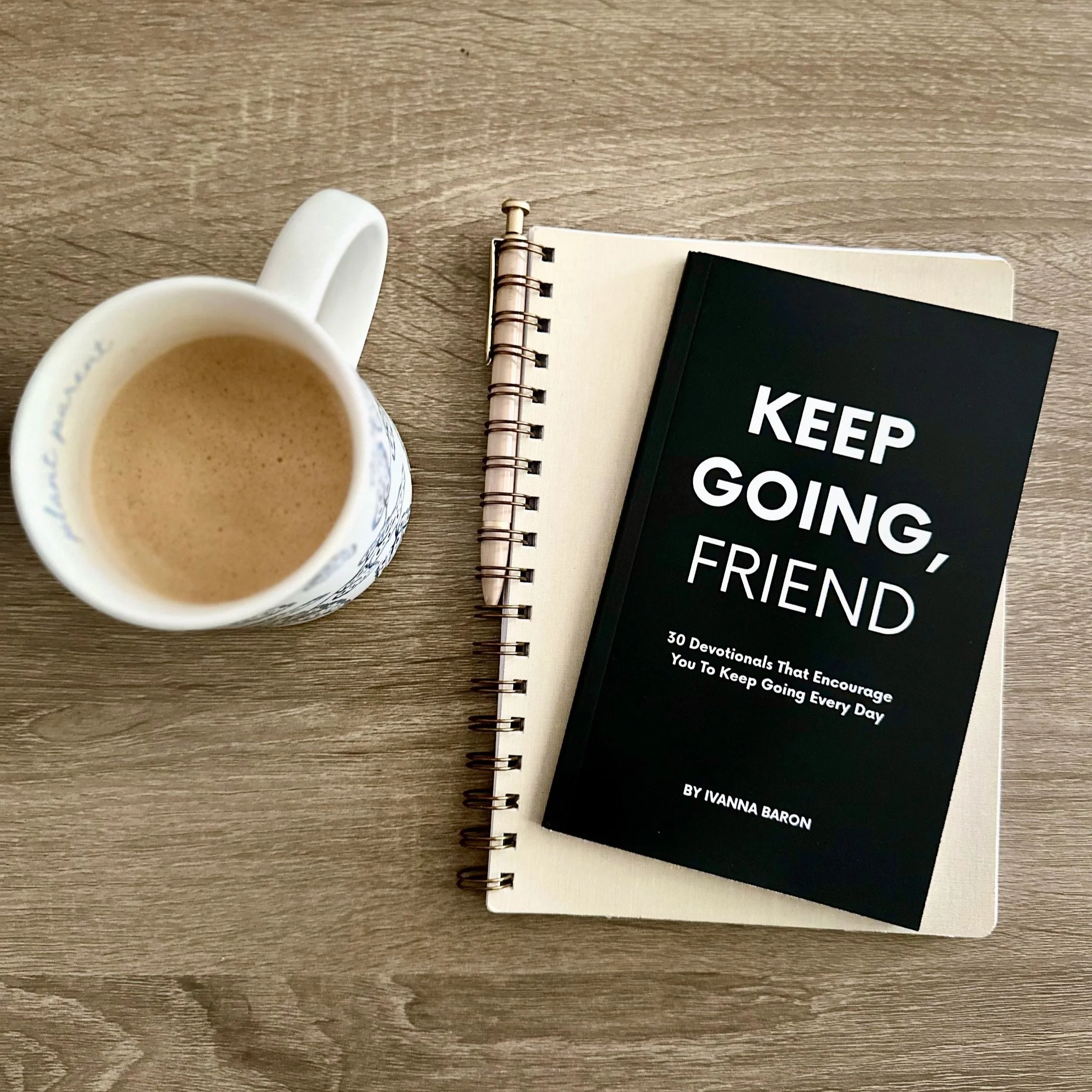 A top-down view of a cup of coffee with a spiral-bound notebook and a book titled 'Keep Going, Friend' on a wooden table.