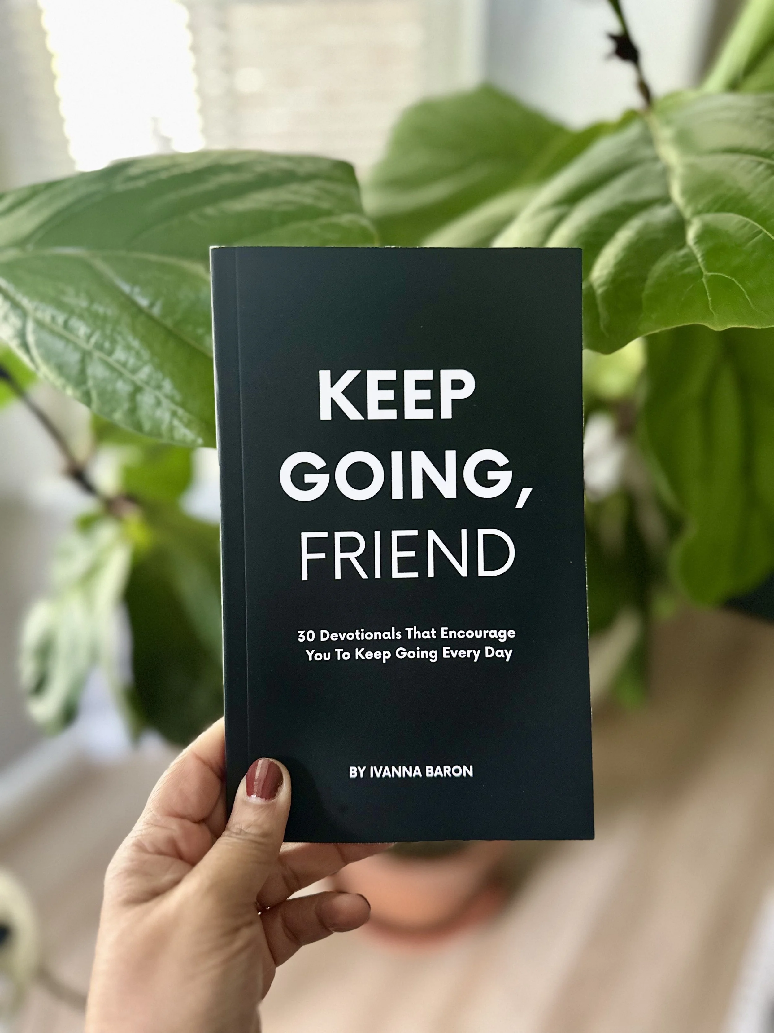 A hand holding a book titled 'KEEP GOING, FRIEND' by Ivanna Baron, with green leaves of a houseplant in the background.