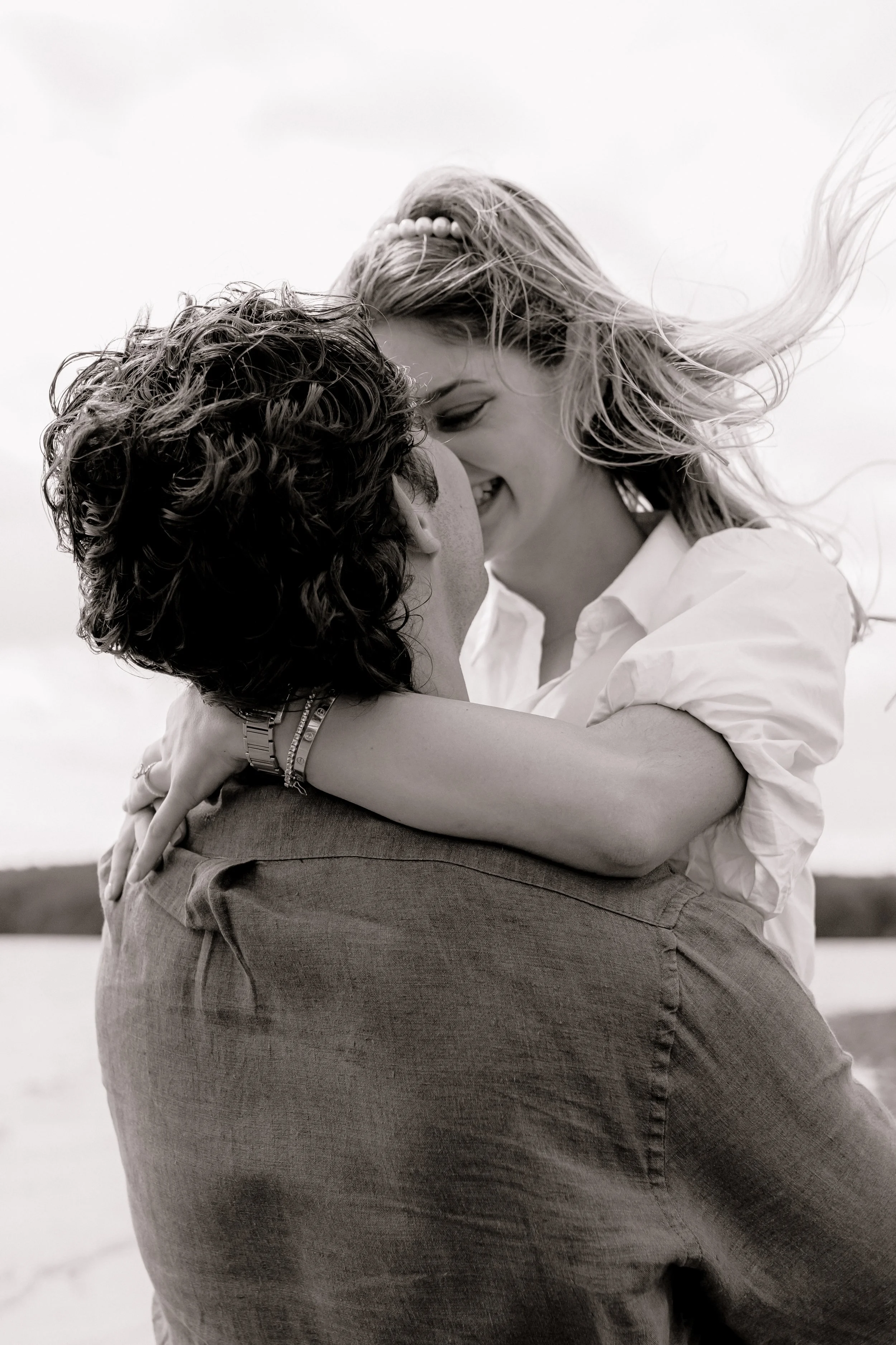 A black-and-white photo of a smiling woman with wind-blown hair embracing and leaning close to a man with curly hair outdoors near a body of water.