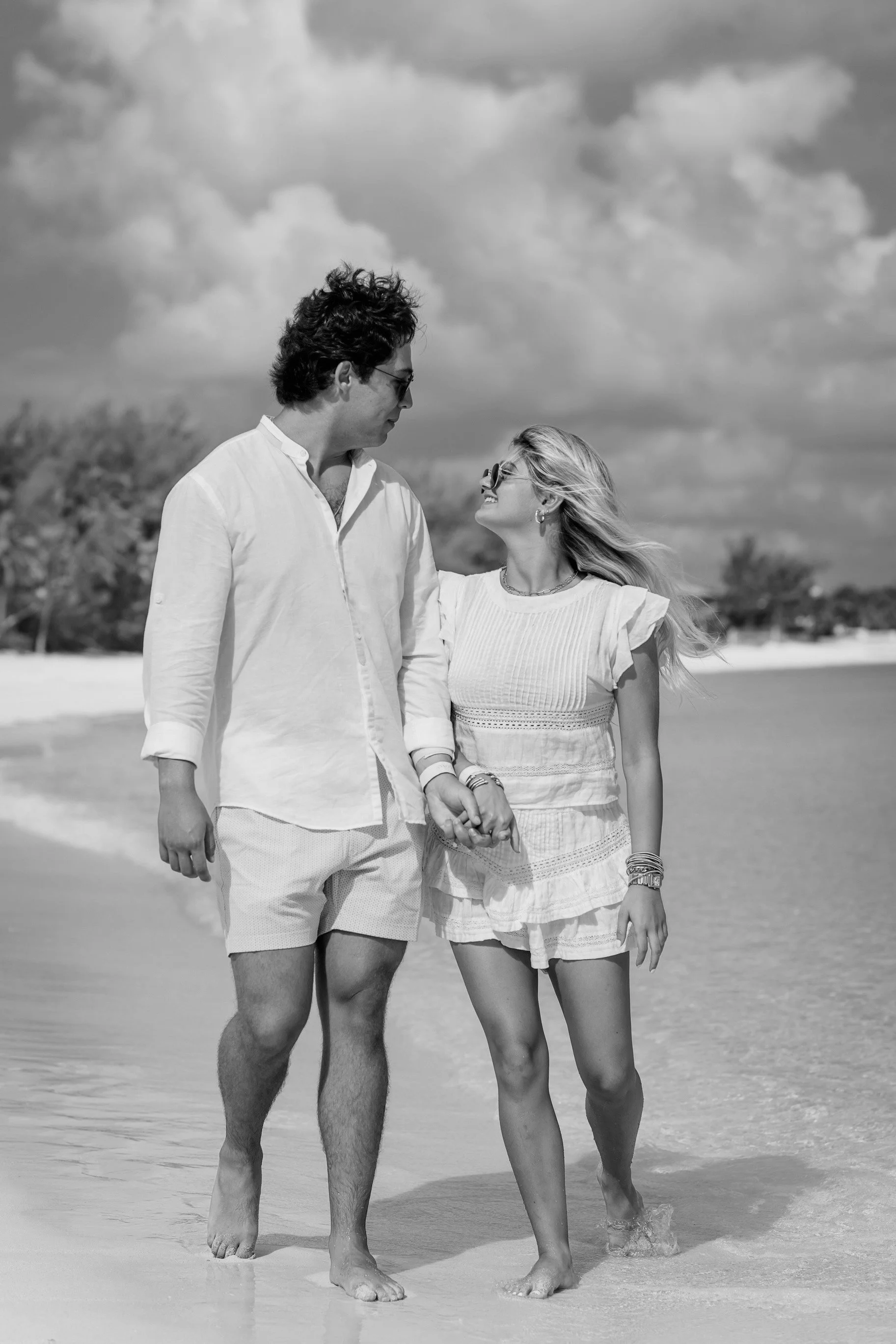 A couple walking hand in hand on a beach, smiling and looking at each other