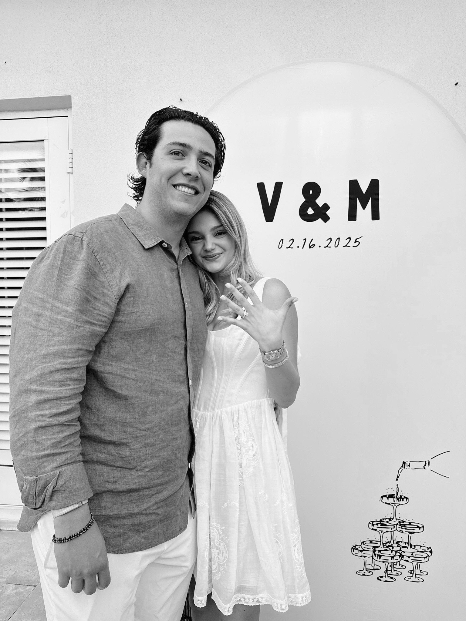 A couple stands together in front of a wall with 'V & M' and a date on it. The woman shows off a ring on her hand.