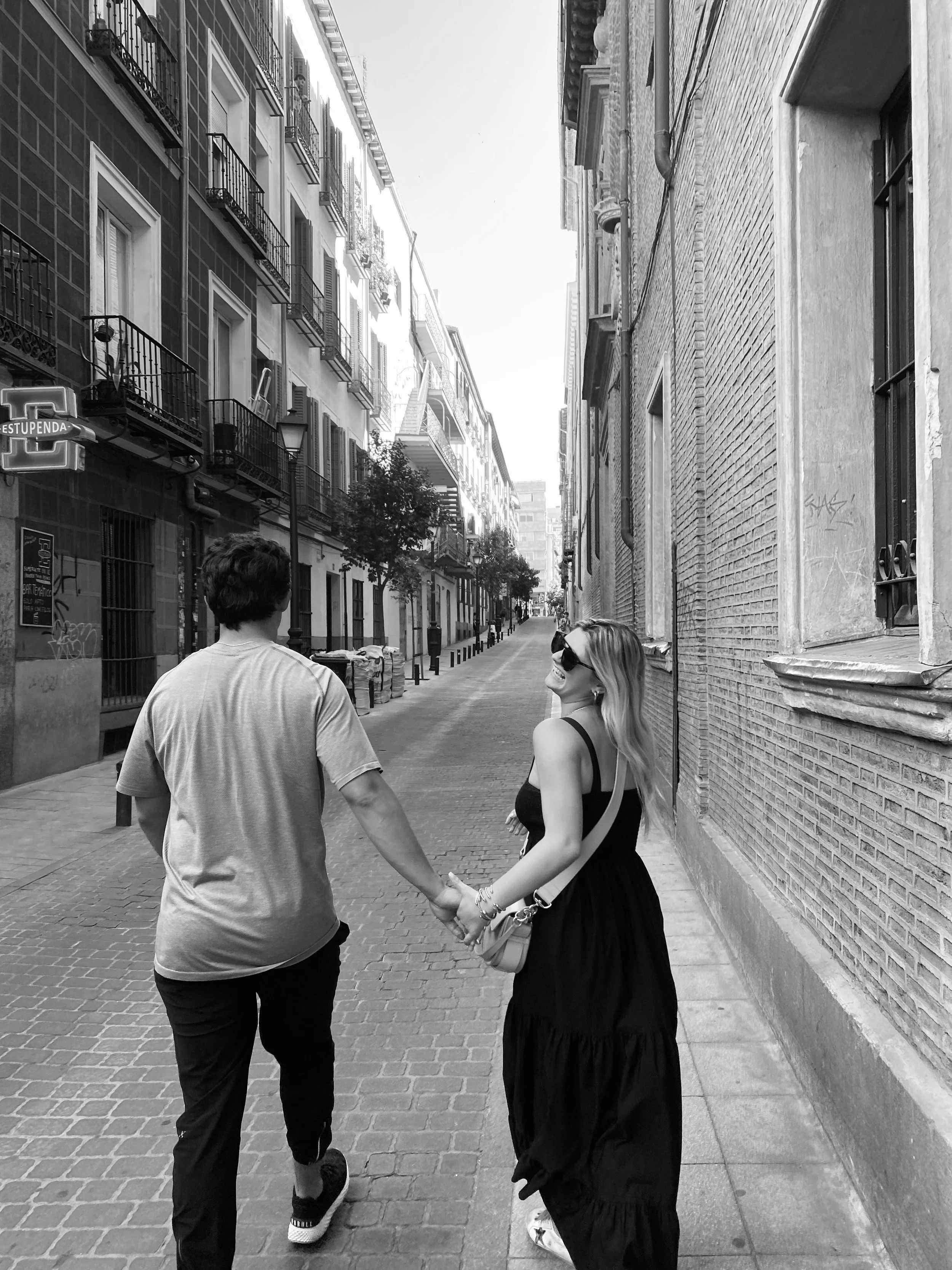 A black and white photo of a couple holding hands and walking down a narrow city street lined with apartment buildings and trees.