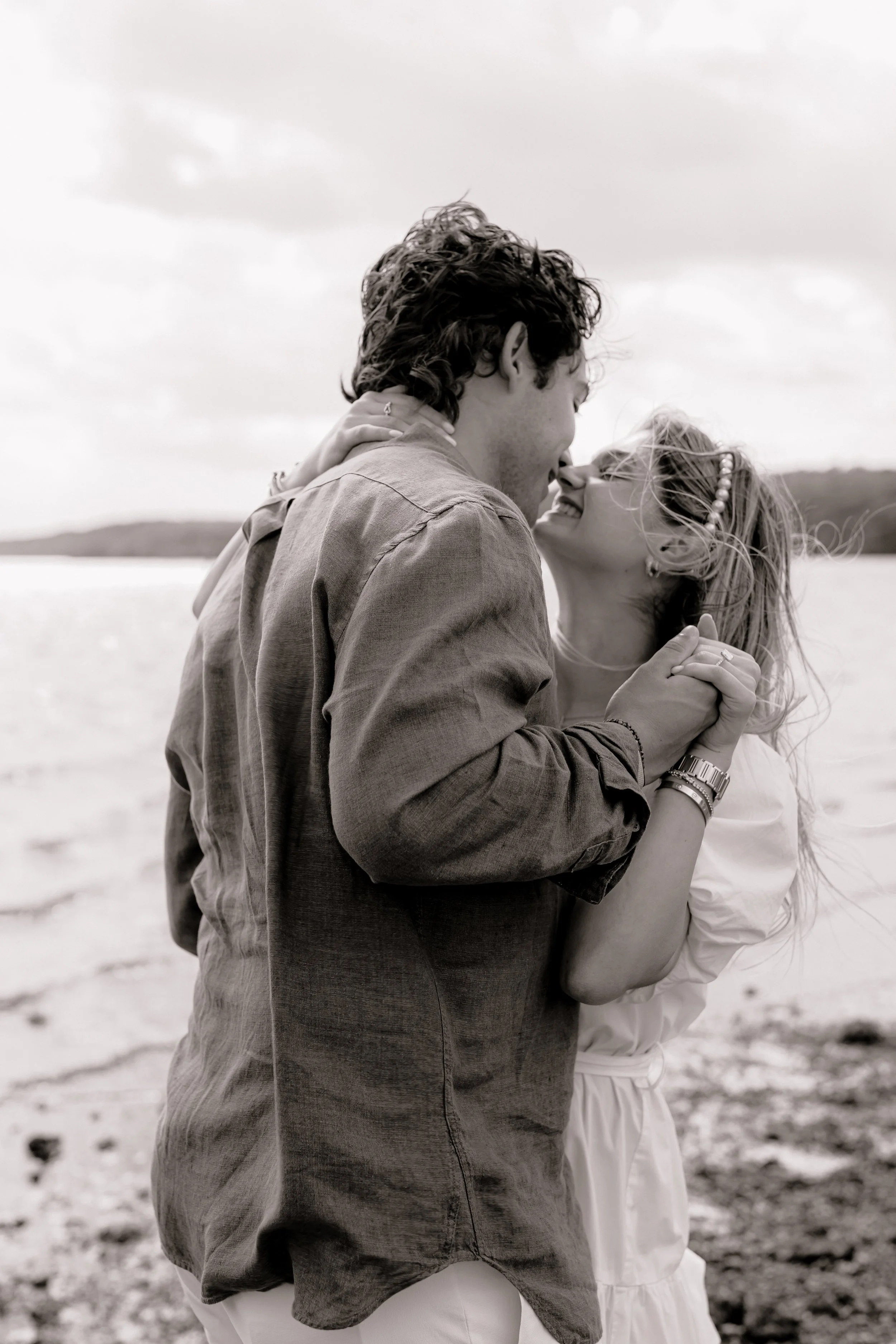 A black and white photo of a young couple holding hands and smiling at each other while dancing on a beach, with water and distant land or trees in the background.