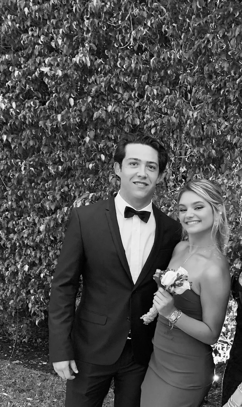 A young man in a tuxedo and a young woman in a strapless dress holding a small bouquet of flowers, standing in front of a leafy background, smiling for a photo.