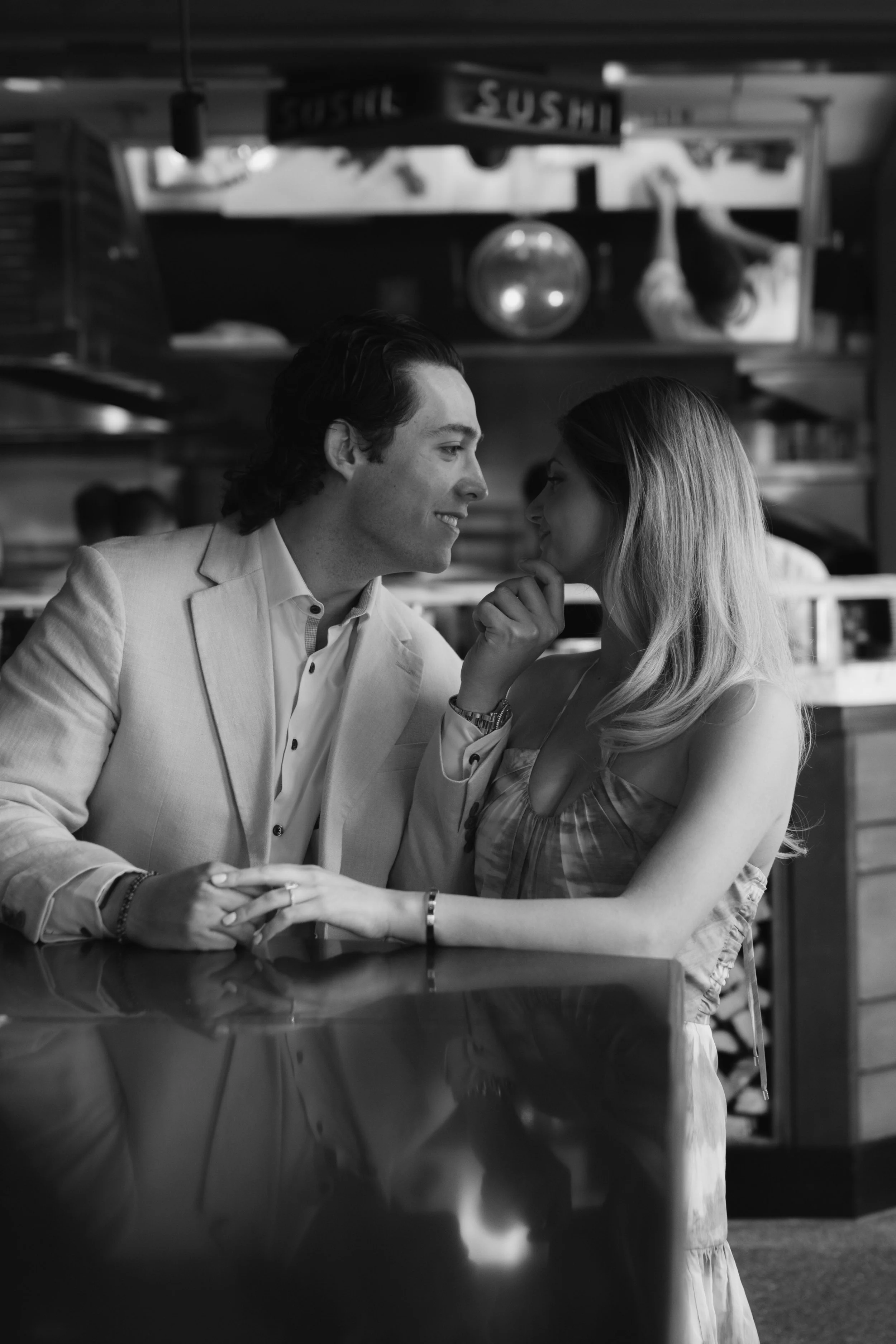 A young couple in an intimate moment sitting at a restaurant, gazing into each other's eyes, with a culinary-themed sign and decor in the background.
