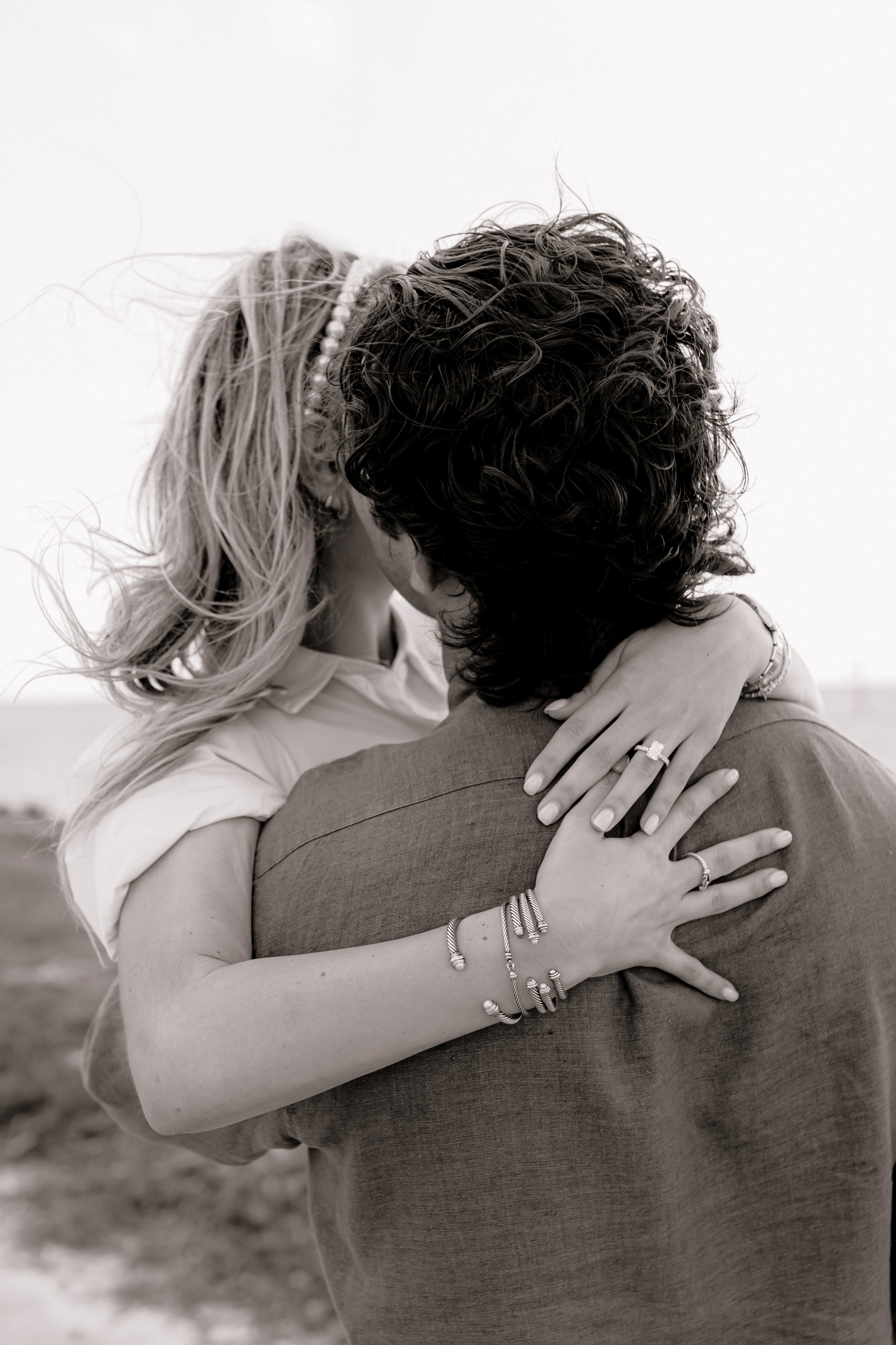 A woman and a man embrace passionately outdoors, with their faces close together and eyes closed. The woman has long, wavy hair, and is wearing bracelets and rings. The man has curly hair and is dressed in a blazer. The image is in black and white.