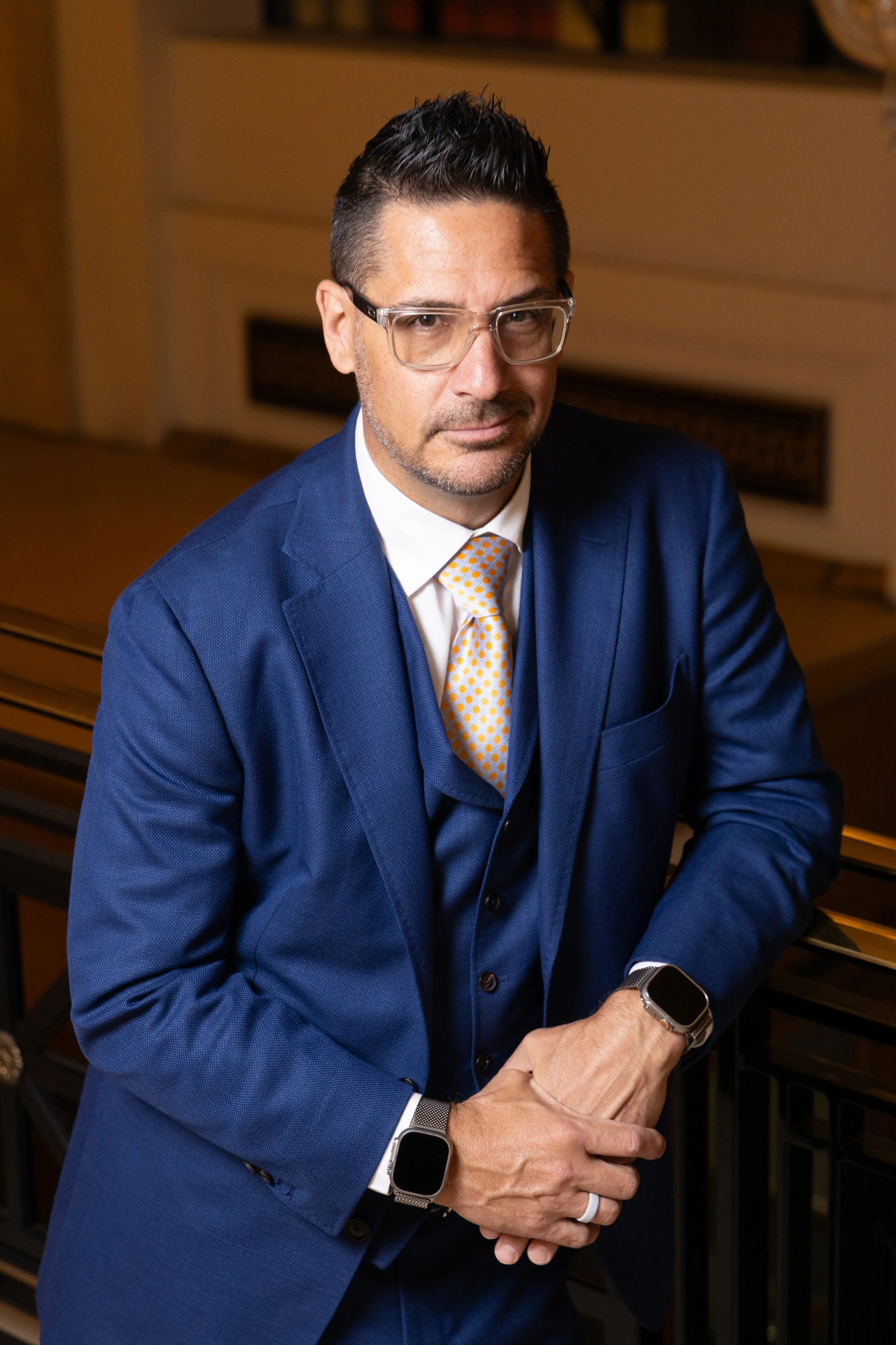 A man with dark hair, glasses, and light facial hair wearing a blue blazer, a white shirt, and a yellow polka dot tie, sitting at a piano with a serious expression.