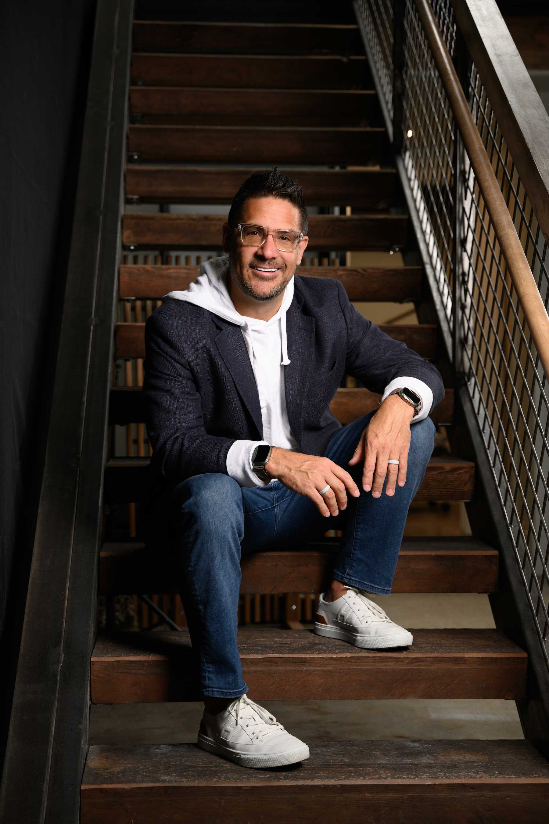 A man sitting on wooden stairs, smiling, wearing glasses, a dark blazer over a hoodie, and white sneakers.