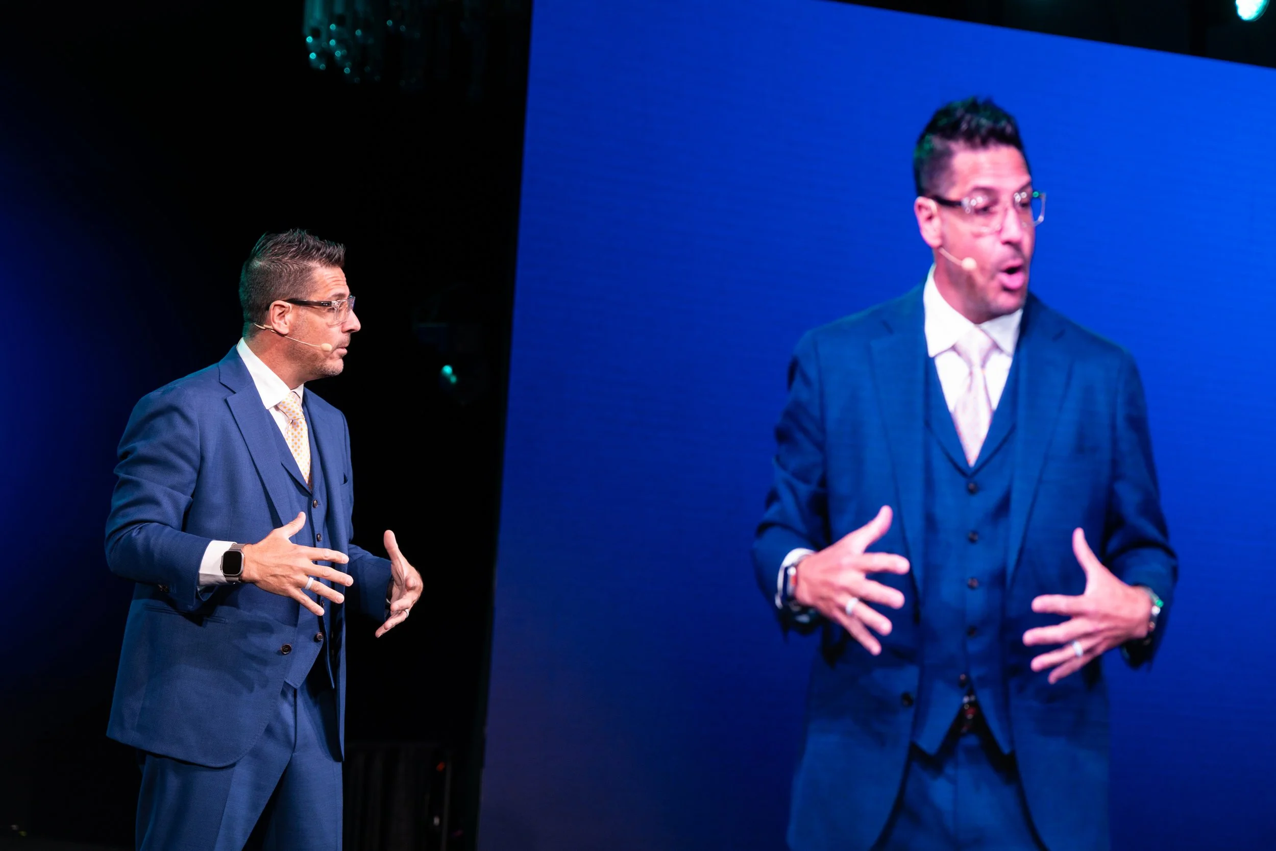 Two men in blue suits speaking on stage with one visible on a large screen behind them. The man on the left is wearing glasses, has short hair, and is gesturing with his hands. The man on the right is also gesturing and speaking into a microphone. The background is dark with stage lighting.