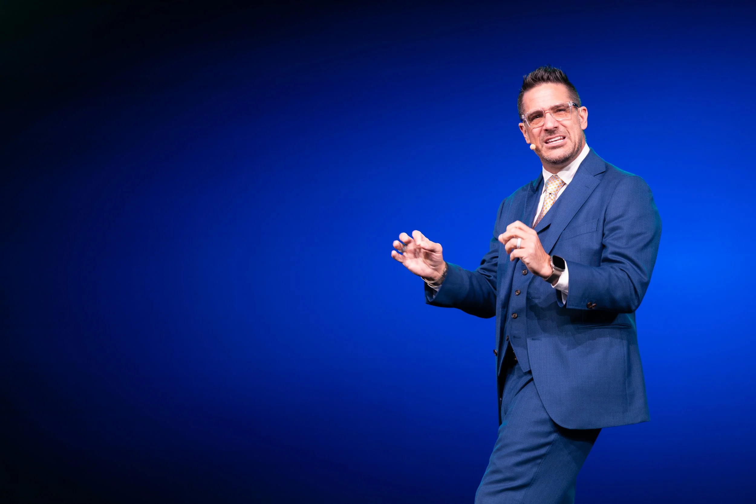 A man wearing a blue suit, glasses, and a yellow tie with white dots, speaking on stage against a blue background.