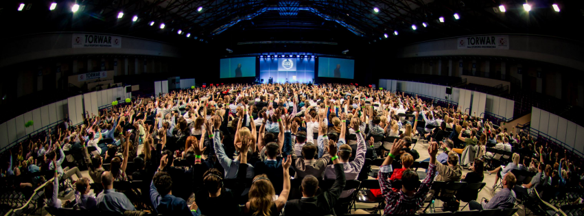 Large conference hall filled with a crowd of people seated at tables, raising their hands, facing a stage with large screens at the front.