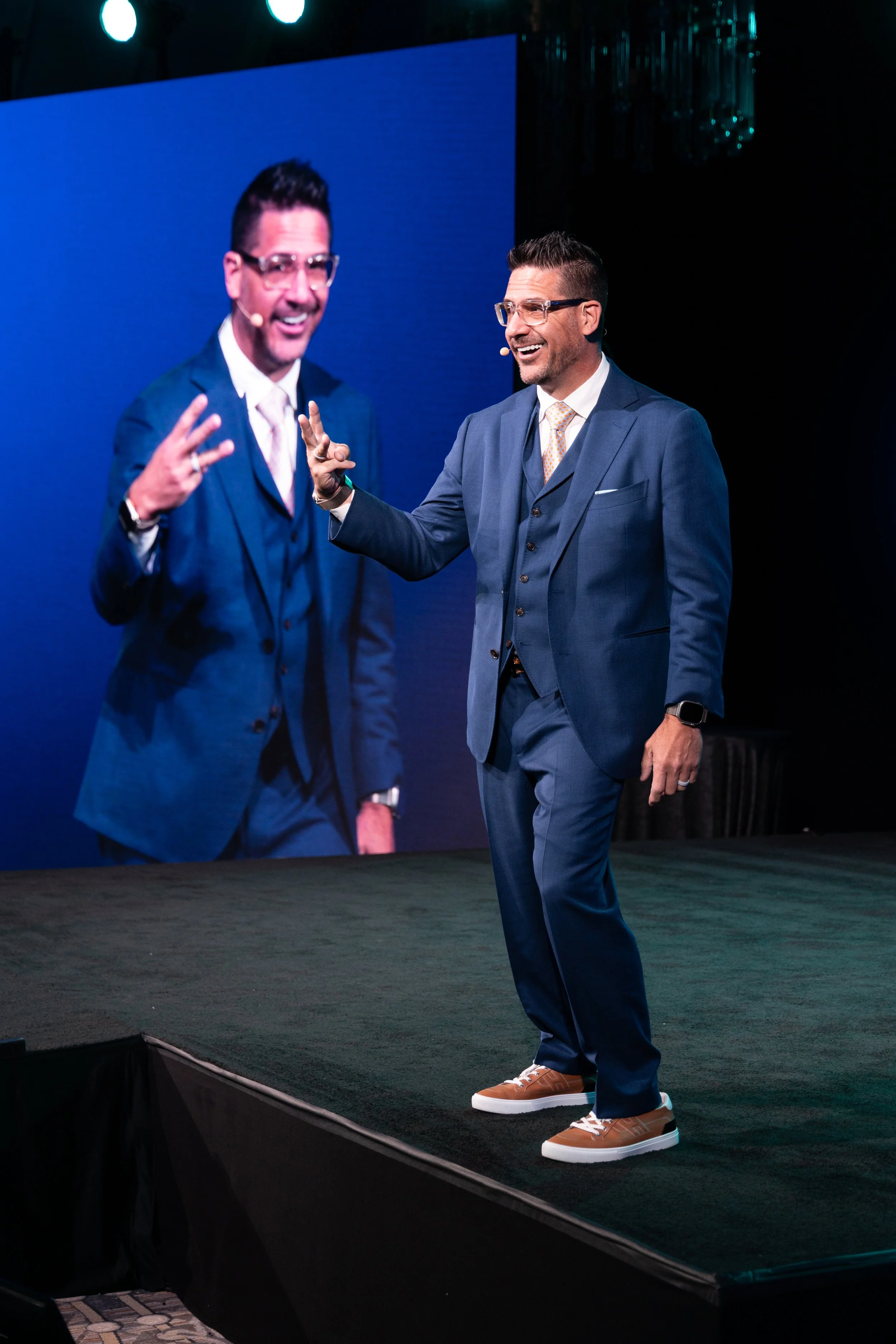 A man in a blue suit and glasses is standing on a stage, smiling and gesturing with his hand. A large screen behind him displays his image. He is wearing brown sneakers with white soles.