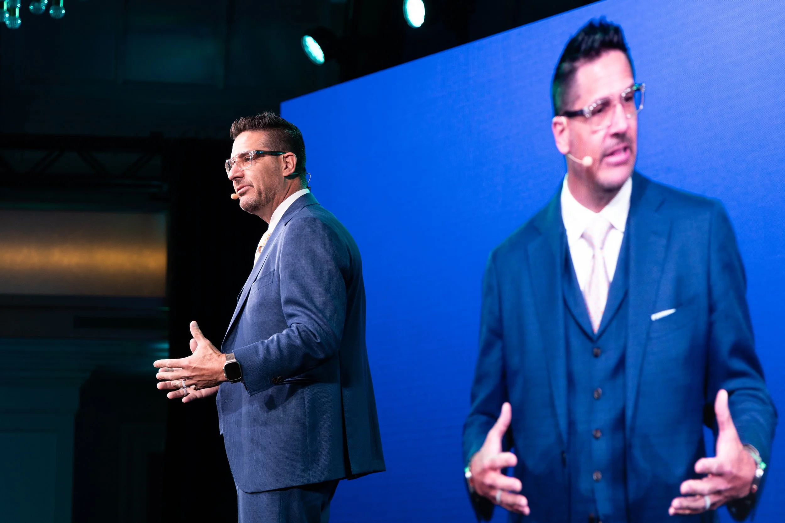 Two men in blue suits speaking on stage during a presentation, with a large screen displaying their image behind them.