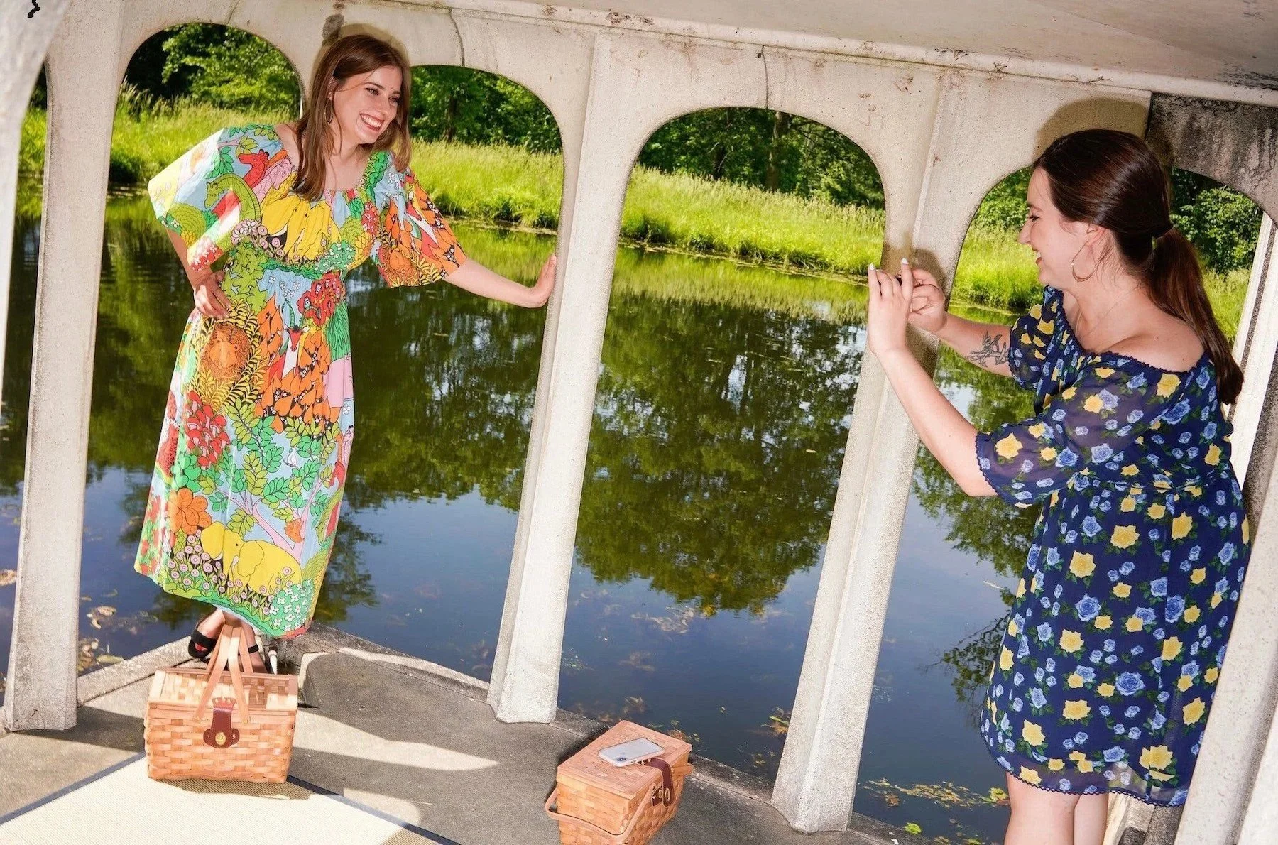 Two women are taking photos of each other through a small bridge with the water and trees in the background. One woman is standing on a picnic basket, wearing a colorful, patterned dress, smiling while posing. The other woman, with a ponytail and tattoos, is in a blue floral dress, holding her phone to take a picture.