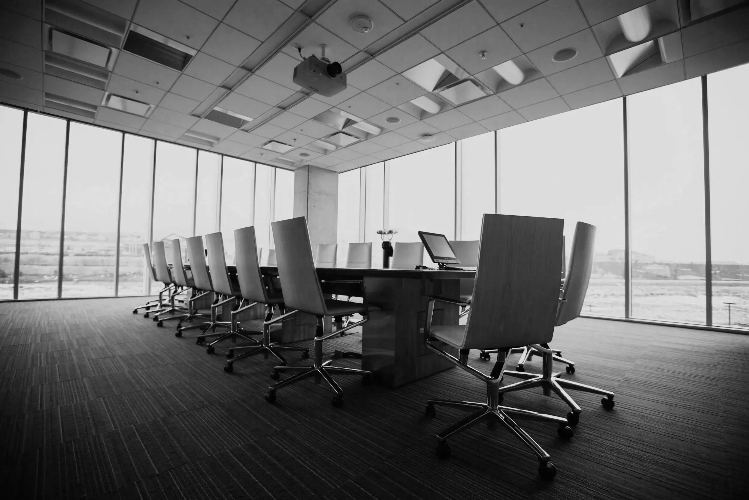 A modern conference room with a long table and multiple chairs, large windows with a city view, and a ceiling-mounted projector.