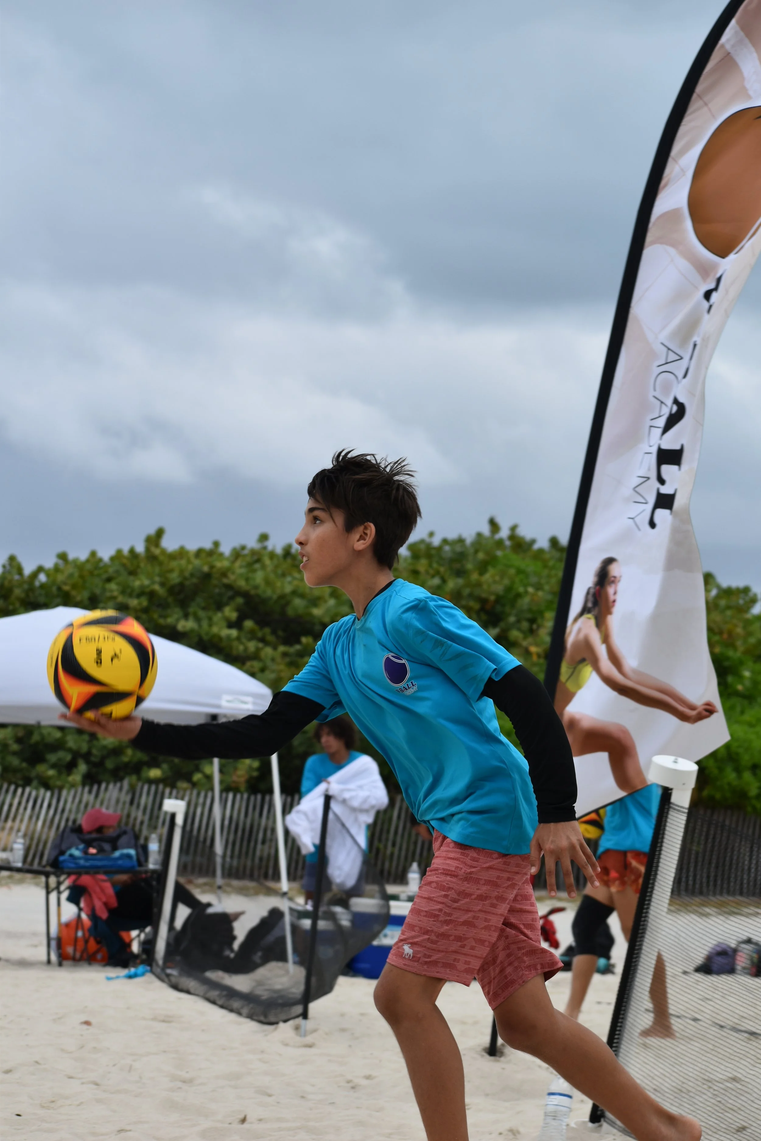 A young boy playing beach volleyball on a sandy beach with cloudy skies, wearing a blue shirt and red shorts, holding a yellow and black volleyball, with a volleyball net and umbrellas in the background.