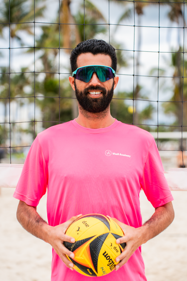 Man standing on a beach volleyball court holding a volleyball, wearing pink T-shirt and sport sunglasses, with palm trees and a volleyball net in the background.