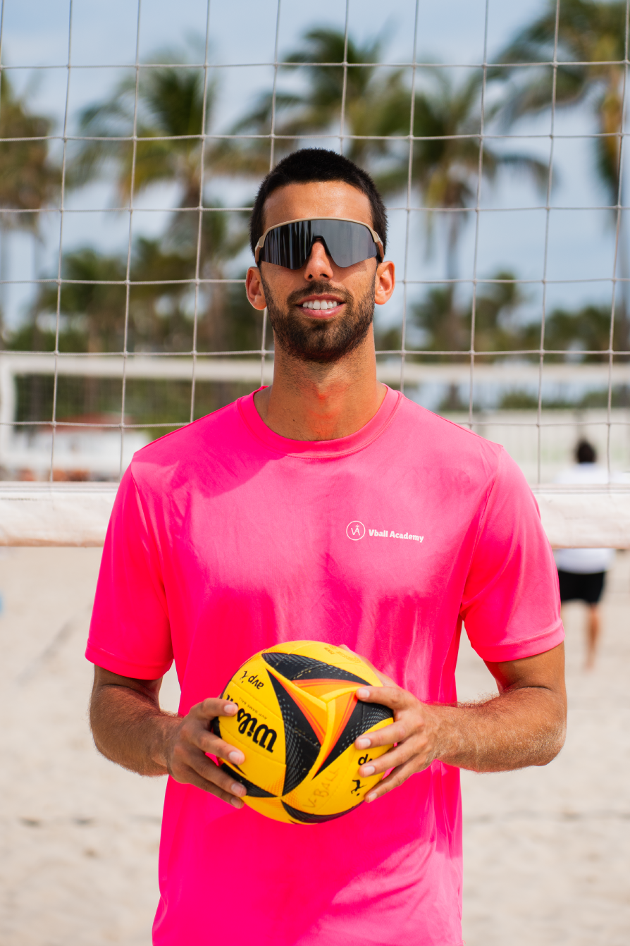 Man holding a yellow and black volleyball on a beach volleyball court, wearing sunglasses and a pink shirt, with palm trees in the background.