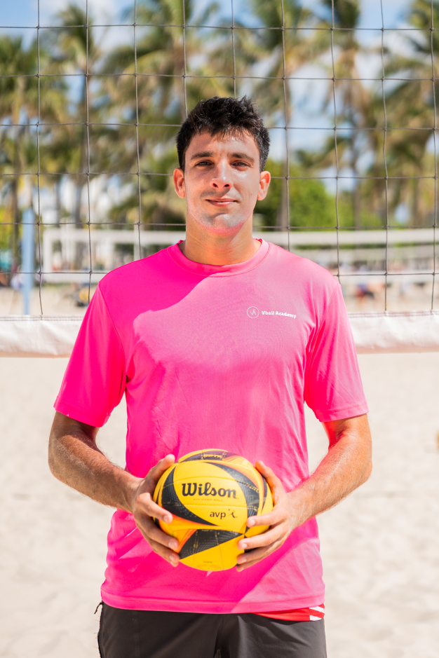 A young man in a bright pink t-shirt holding a yellow and black volleyball on a beach volleyball court, with palm trees and a volleyball net in the background.