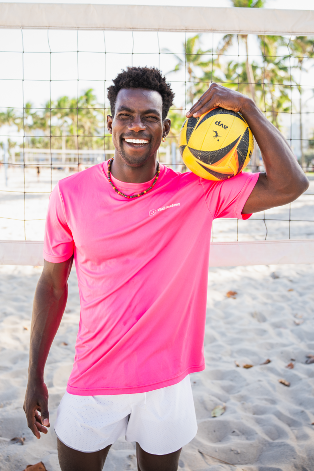A man smiling and holding a yellow and black volleyball on his shoulder, standing on a sandy beach volleyball court with a net and palm trees in the background.