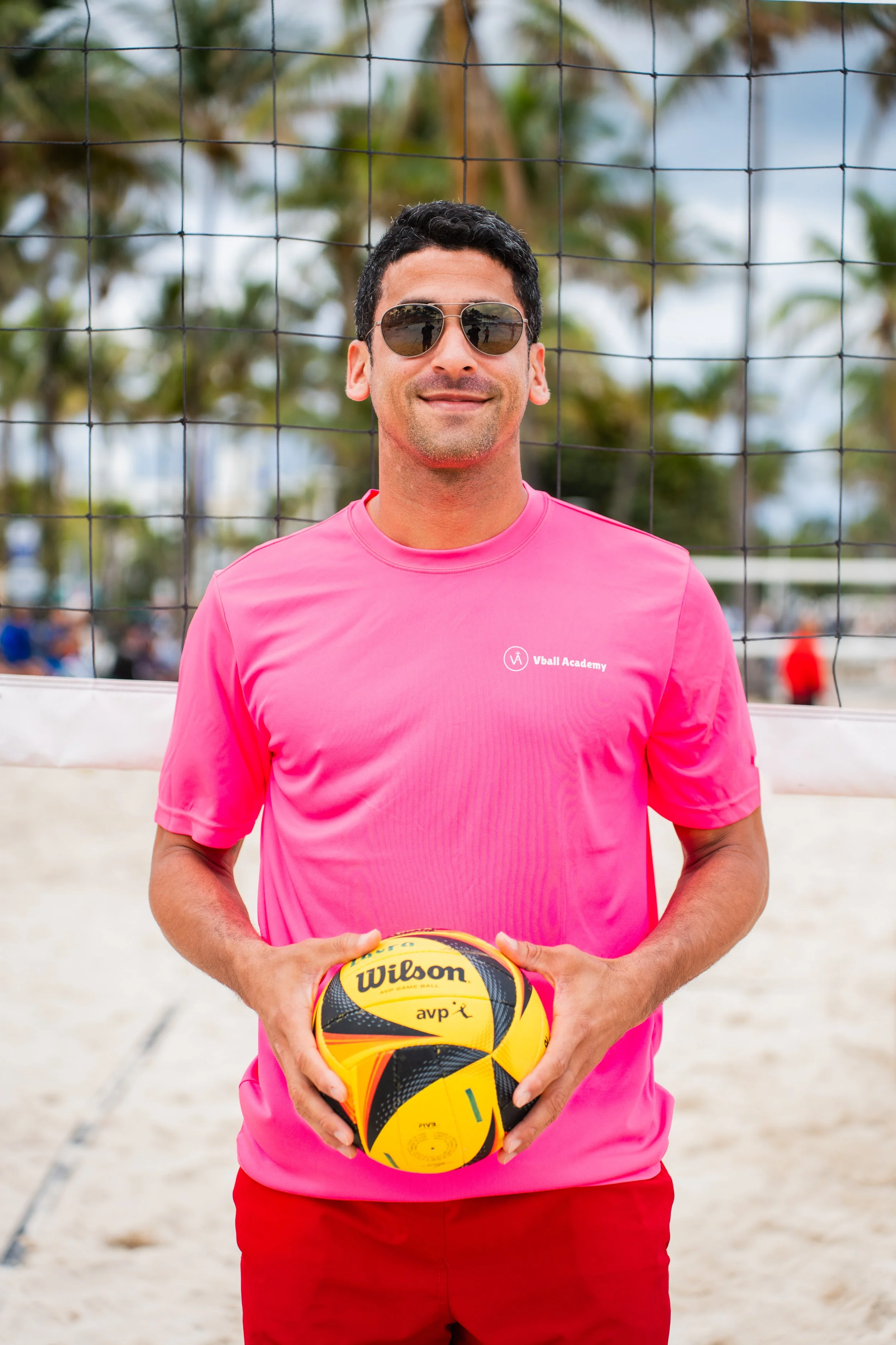 Man holding a yellow and black Wilson volleyball on a beach volleyball court.