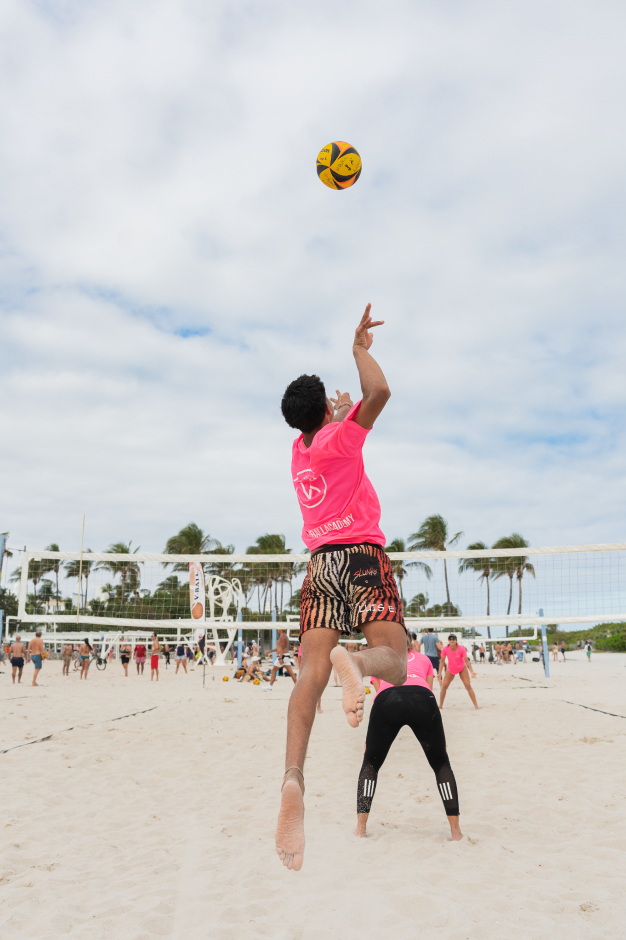 Person jumping to hit a volleyball on a beach volleyball court, with other players and palm trees in the background.