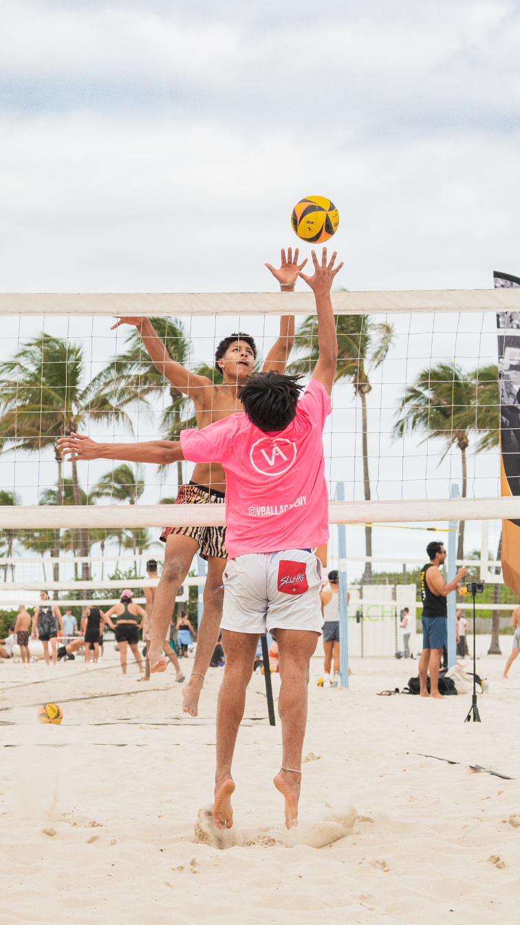 Two men playing beach volleyball, one jumping near the net and the other hitting the ball with his hand. Palm trees and other players are in the background, along with overcast skies.