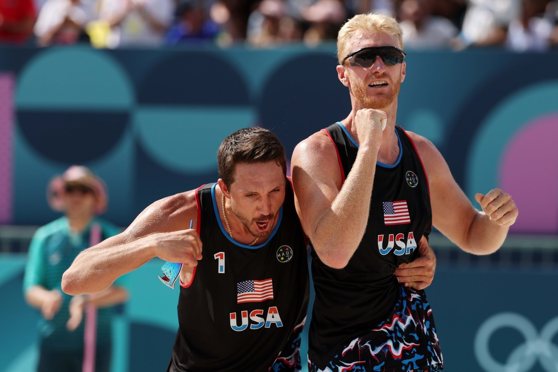 Two male beach volleyball players from the USA celebrating during a match, with a crowd in the background, wearing black jerseys with patriotic USA flags and American flags on their shorts.