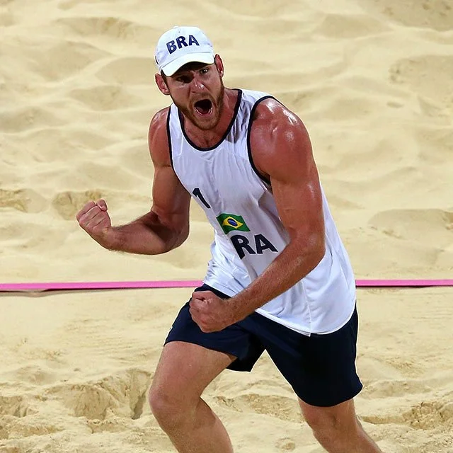 A male beach volleyball player celebrating passionately on the sand, wearing a white cap with 'BRA' written on it, a white sleeveless jersey with Brazilian flag, and black shorts.