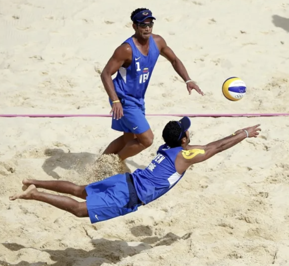 Two male beach volleyball players in blue uniforms are diving and reaching for a volleyball on sandy court during a match.
