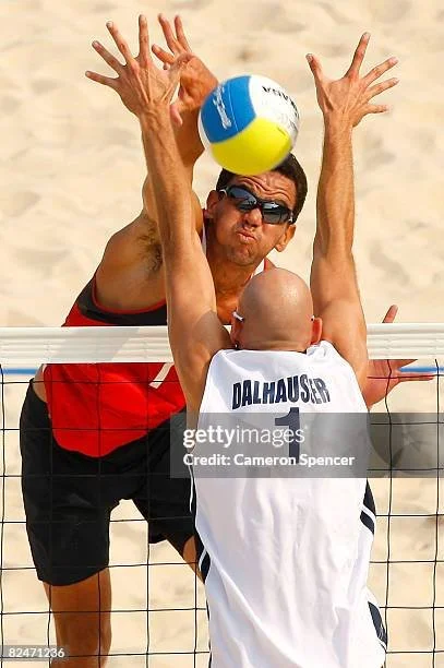 Two men playing beach volleyball, one with sunglasses hitting the ball over the net, the other with a shaved head attempting to block.