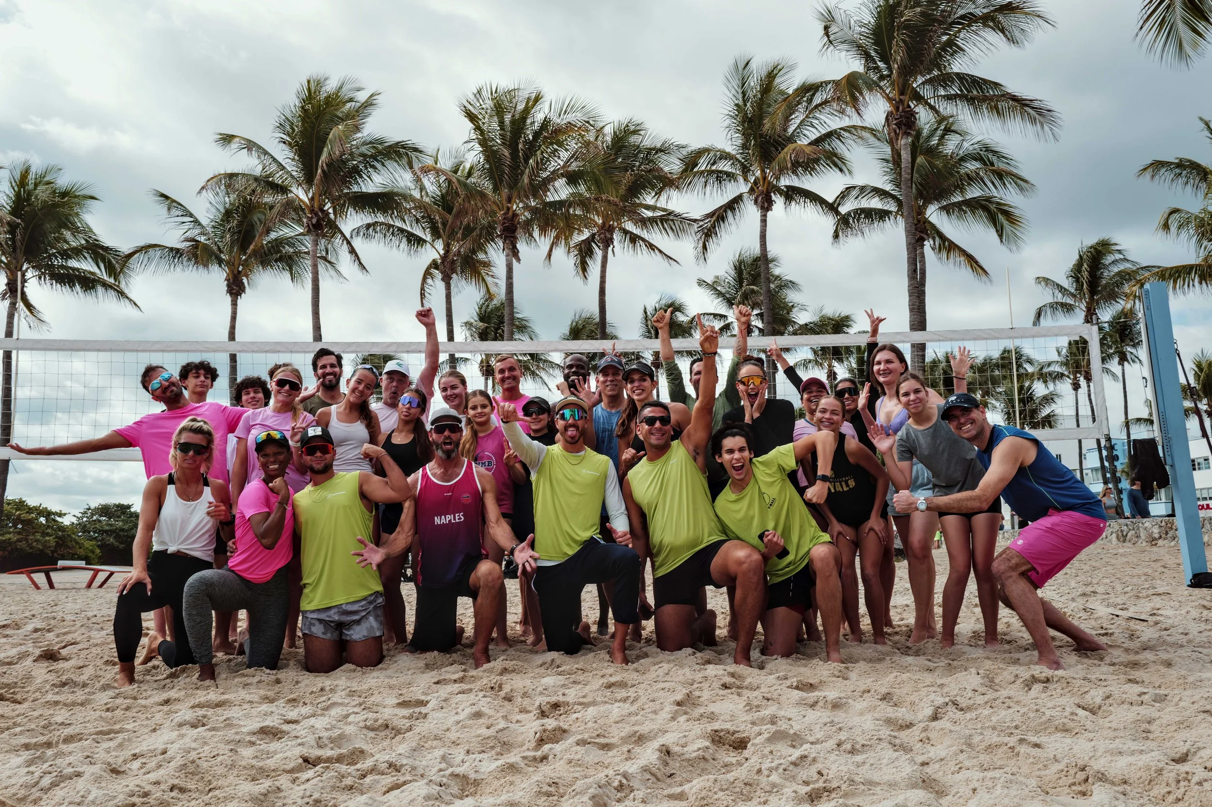 Group of people celebrating on a sandy beach in front of a volleyball net, with palm trees and cloudy sky in the background.